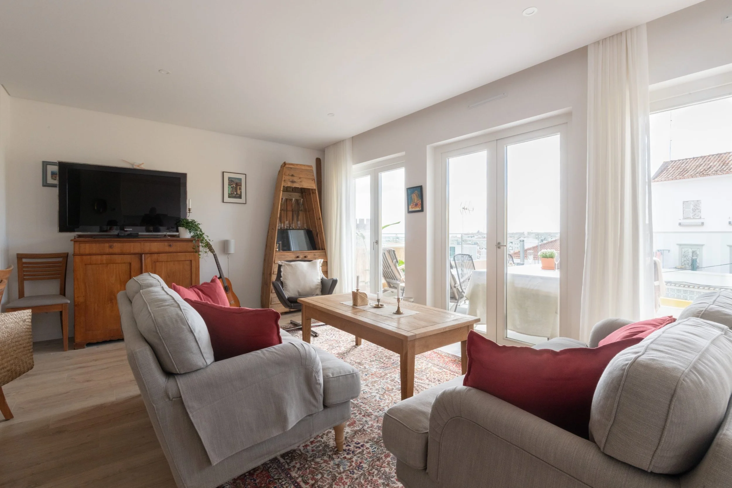 Living room with beige sofas, red pillows, wooden coffee table, TV on wooden cabinet, and large glass doors leading to balcony with outdoor furniture and city view, white curtains, wooden flooring.