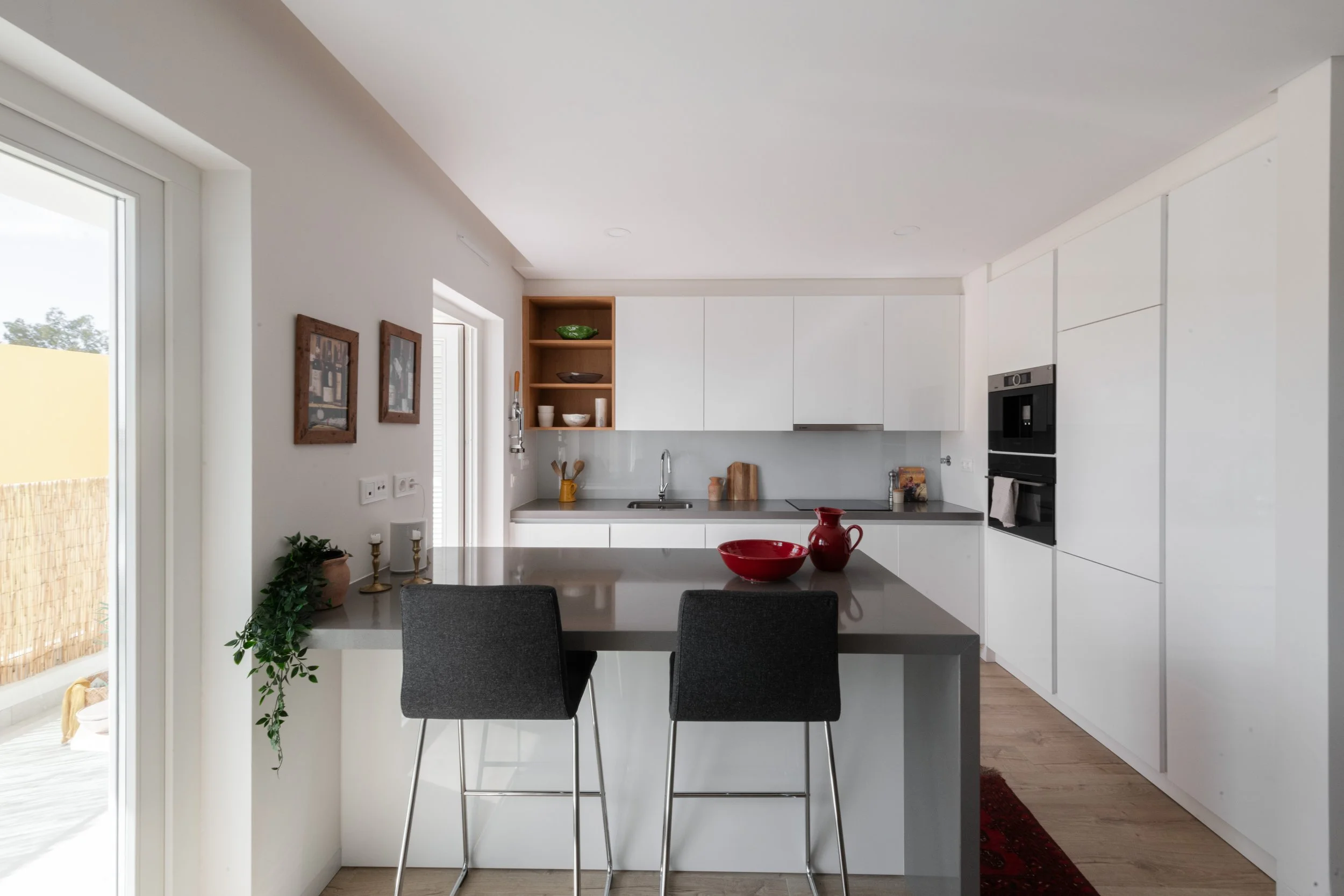 Modern kitchen with white cabinets, gray countertops, and a breakfast bar with two black chairs. Red bowls and pitcher on the counter. Open shelving with bowls and decorative items, and a view of a balcony through glass doors.