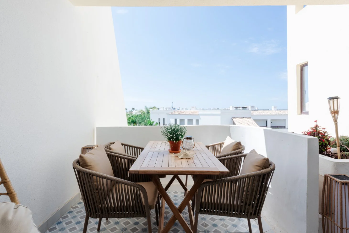 Outdoor balcony with a wooden table, six wicker chairs with pillows, potted plant, lantern, and decorative items, overlooking a white building and blue sky.