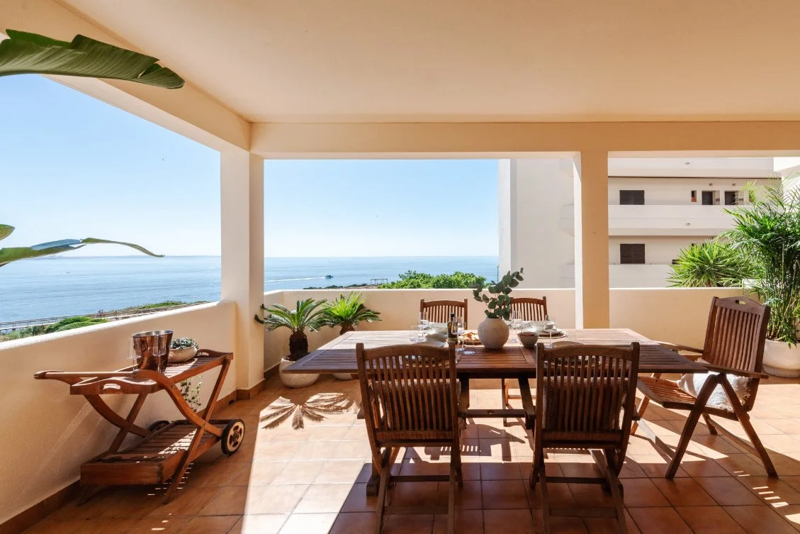 Balcony with wooden dining table and chairs, overlooking the ocean with clear blue sky, potted plants, and a cart on the side.