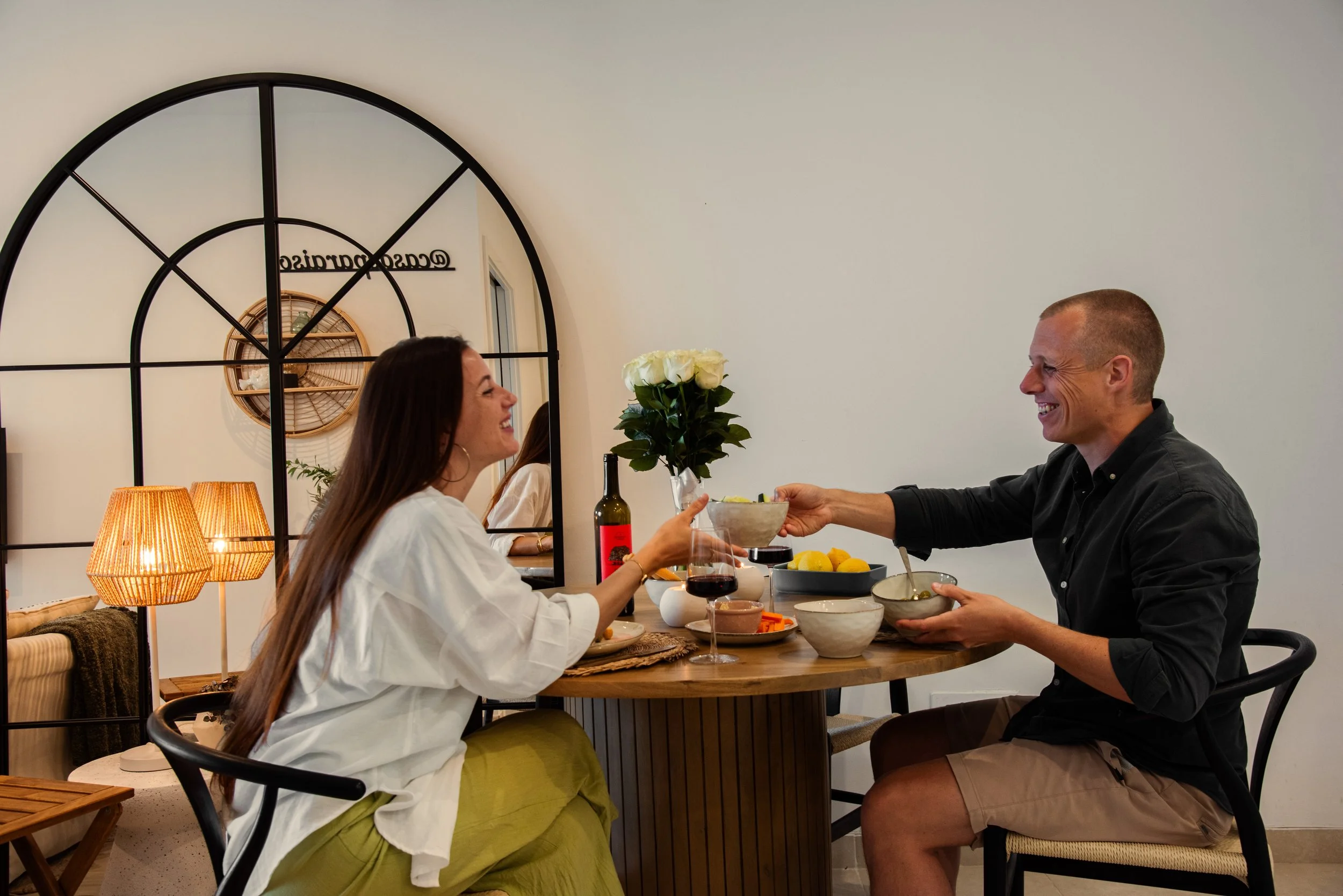 A smiling couple having dinner at a table with wine, bowls, and a bouquet of white roses, in a cozy room with warm lighting and a decorative mirror. Holiday home in Lagos Algarve