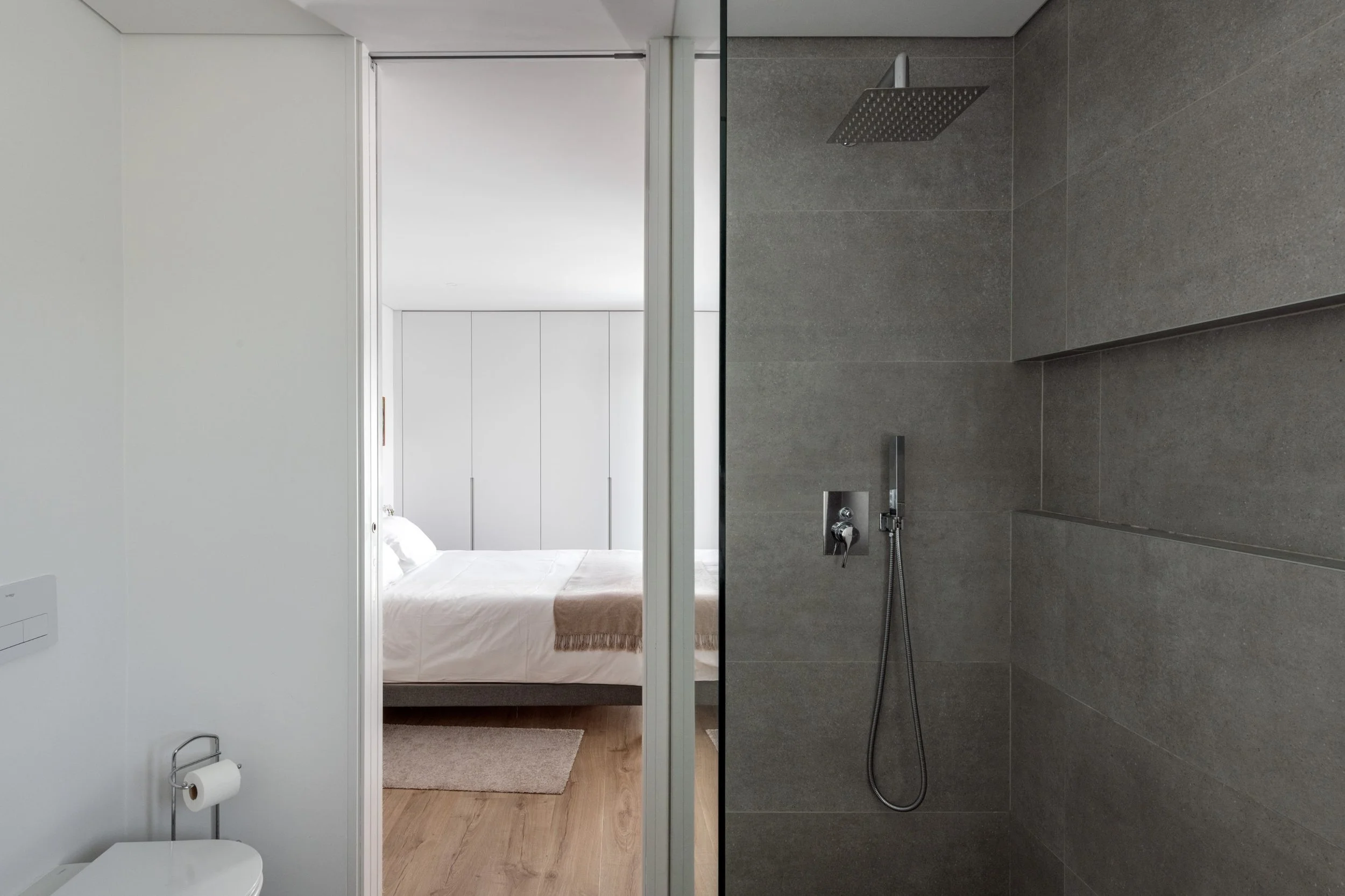 A bedroom visible through a doorway from a bathroom. The bedroom has a bed, white walls, a beige blanket, and a wardrobe, with wooden flooring. The bathroom has a shower area with grey tiles, a rainfall showerhead, and a handheld shower.