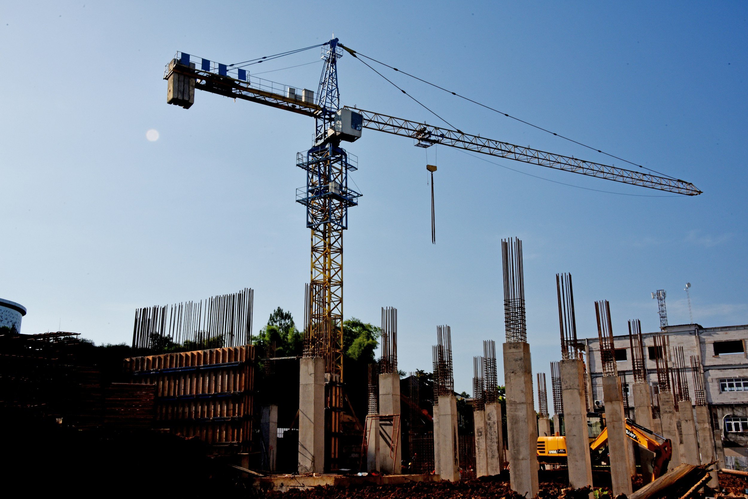 Construction site with a tall yellow tower crane and concrete columns being built, with construction equipment and buildings in the background.