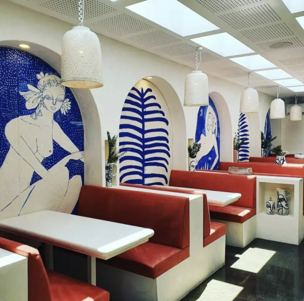 Interior of a restaurant with white walls decorated with blue and white mural art, featuring a woman and stylized leaves, red cushioned booths, white tables, hanging white pendant lights, and potted plants.