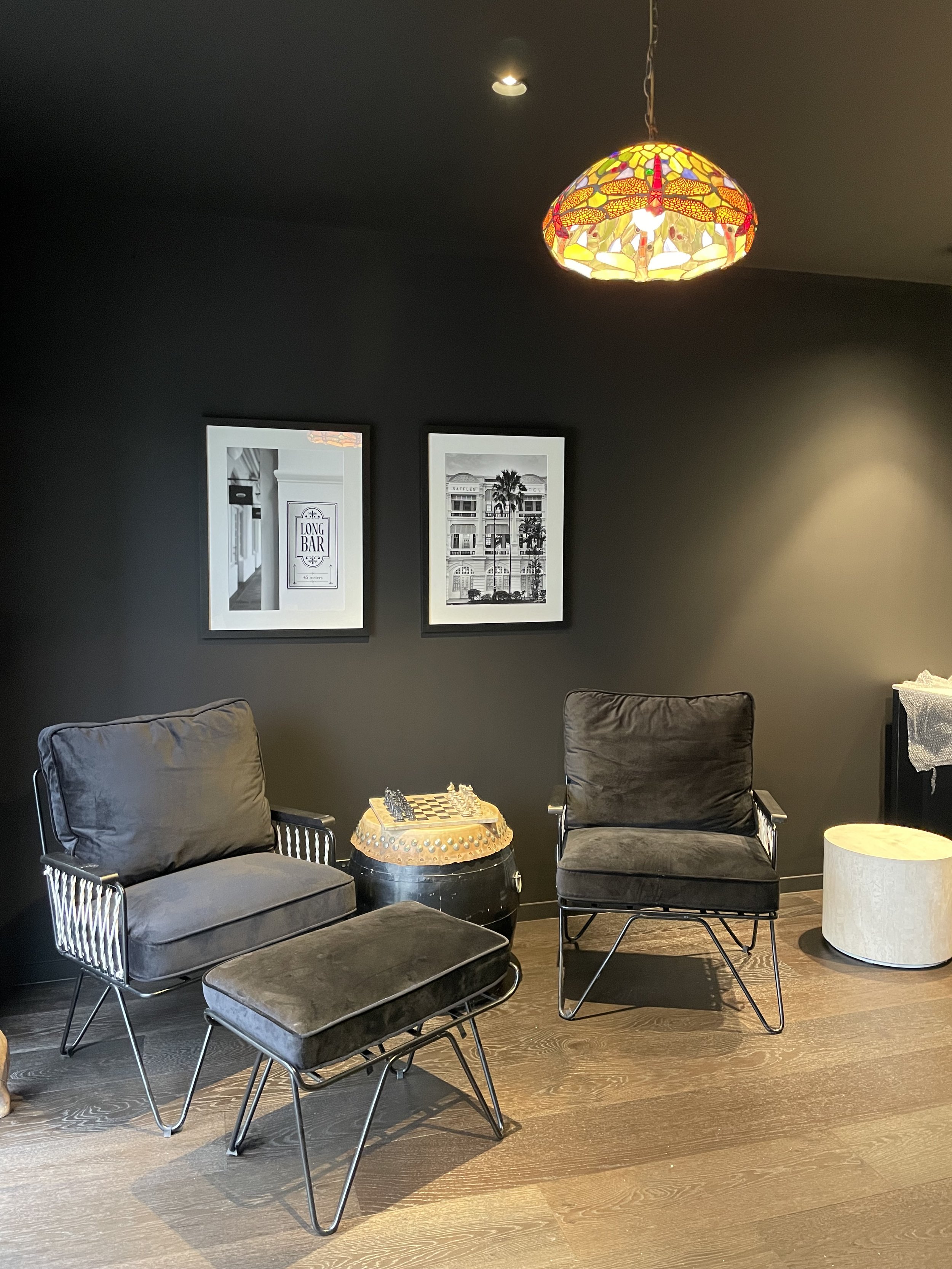 A cozy seating area with two black armchairs, a matching ottoman, a side table with a chess set, a white round side table, artwork on dark gray walls, and a colorful stained-glass pendant lamp hanging from the ceiling.