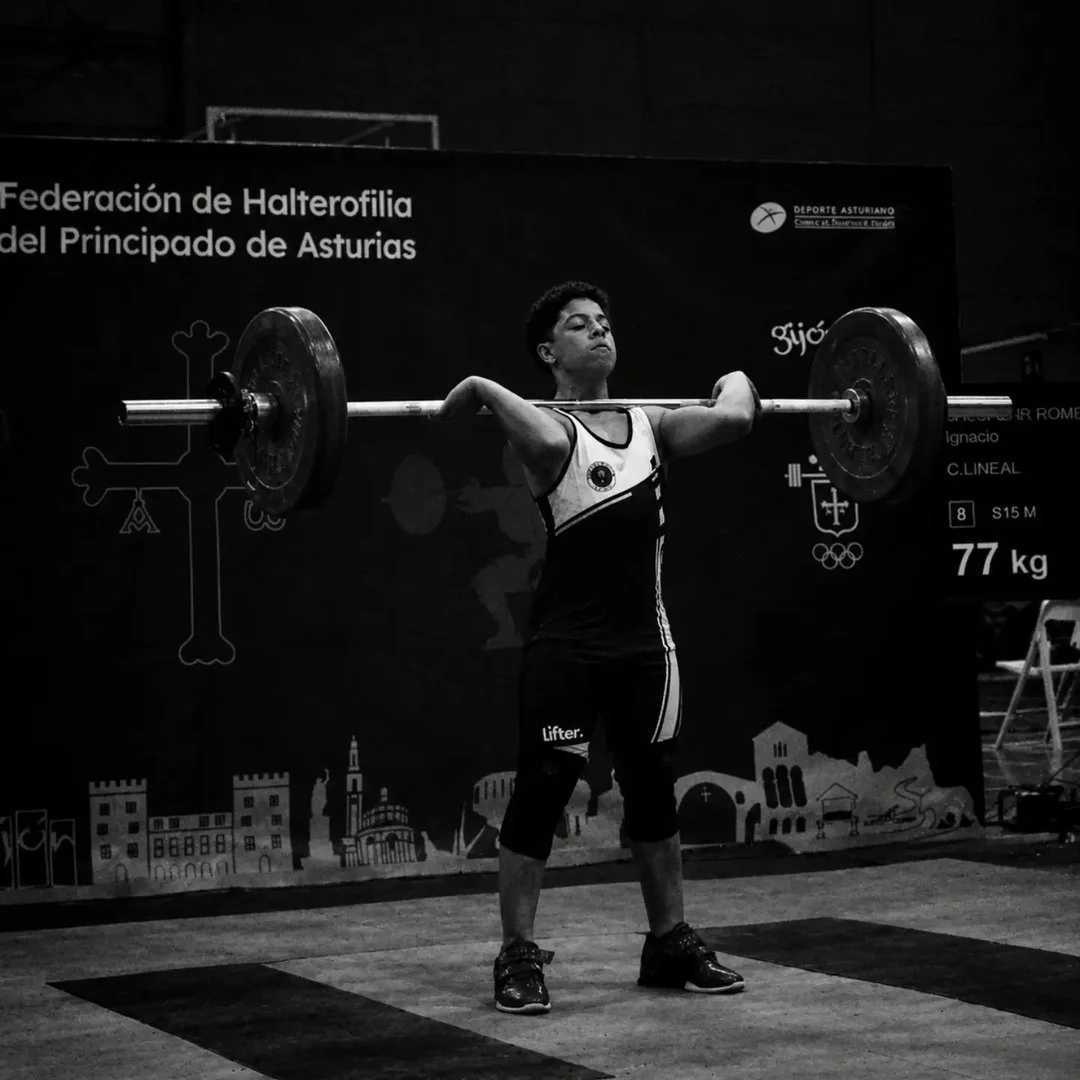 Joven levantando una barra con pesas en una competencia de halterofilia en Asturias, España, en un ambiente de gimnasio o evento deportivo, en blanco y negro.