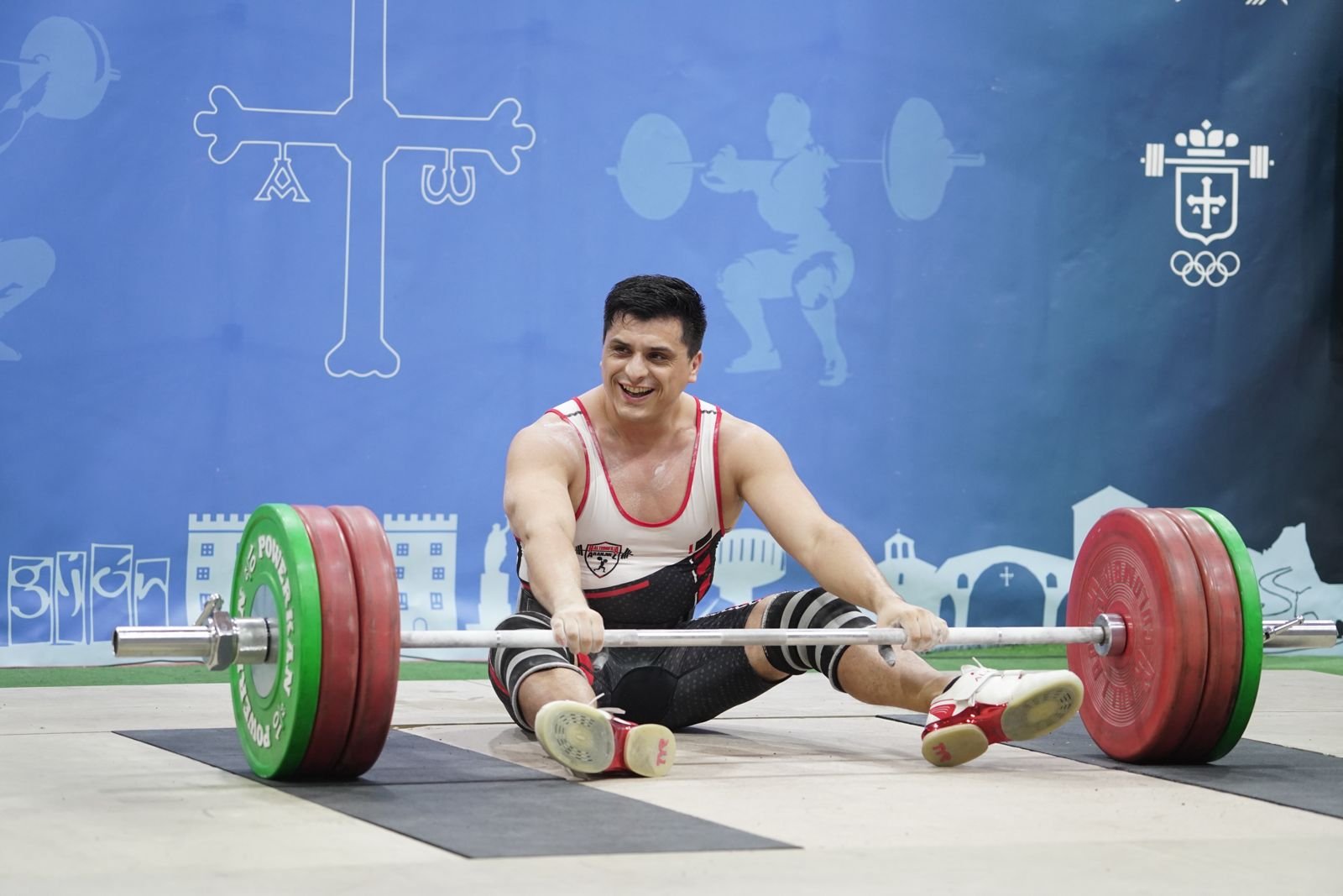Atleta levantando pesas en el suelo durante una competencia, con fondo azul que tiene ilustraciones de deportes y logotipo olímpico.