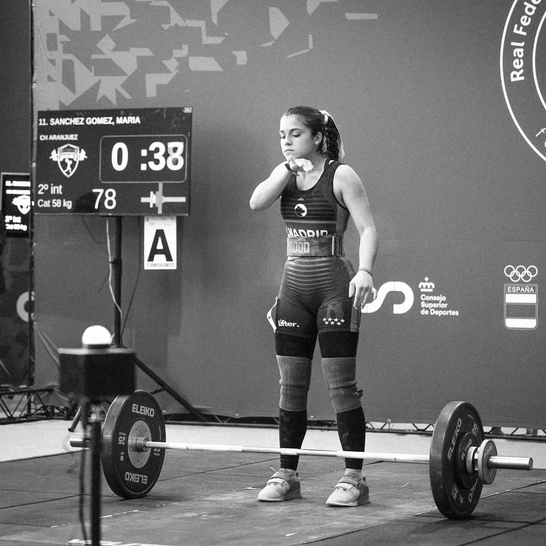 Una joven levantadora de pesas se prepara para levantar una barra con pesas en un entrenamiento o competencia. Ella está concentrada y viste ropa deportiva con el nombre de Madrid. En el fondo hay una pizarra con información y un logotipo del Consejo Superior de Deportes de España.