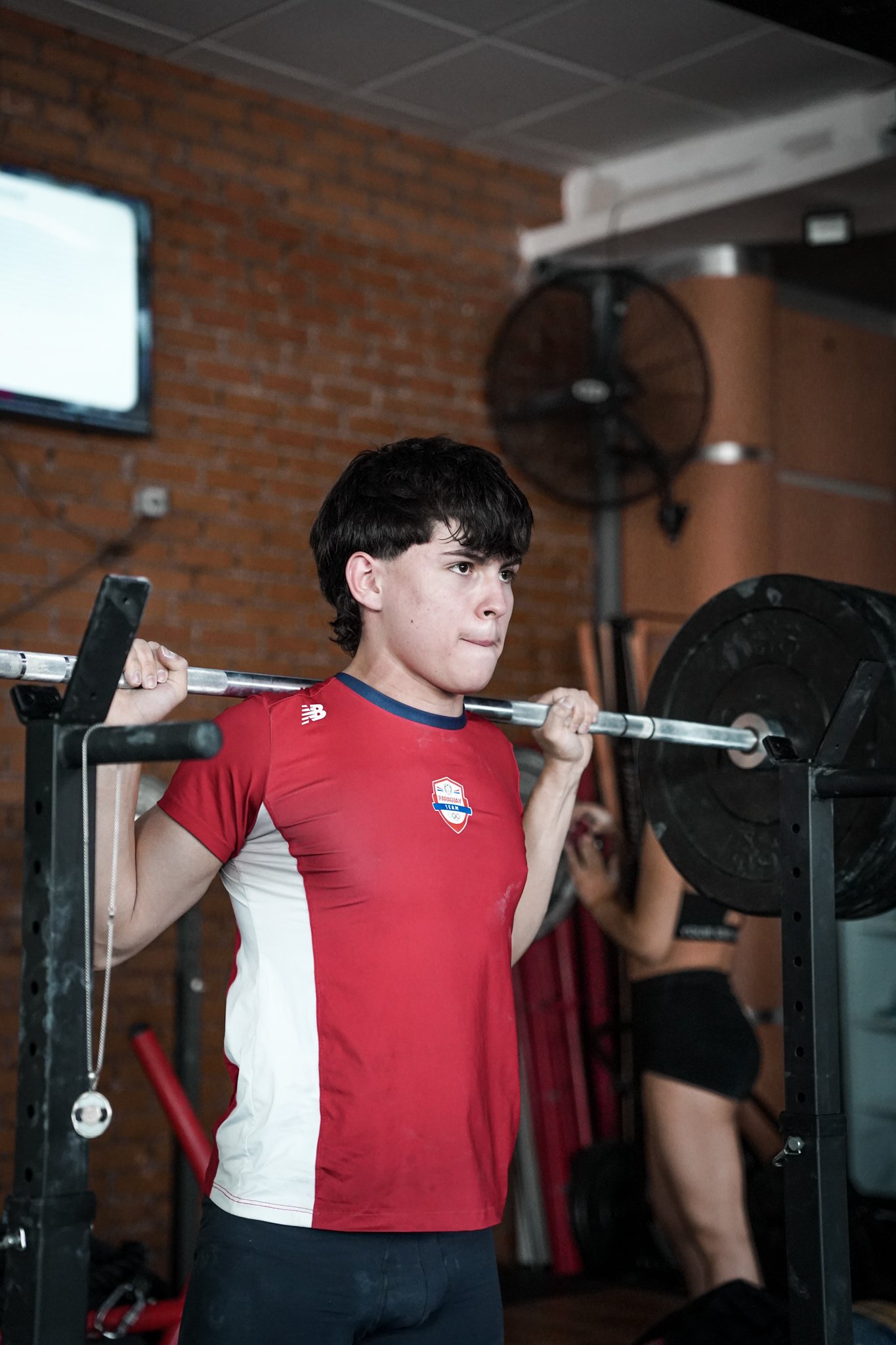 Un joven haciendo sentadillas con barra en un gimnasio. Tiene el cabello oscuro y lleva puesta una camiseta deportiva roja con detalles blancos y un logotipo en el pecho. El fondo muestra una pared de ladrillos y un ventilador de pie.