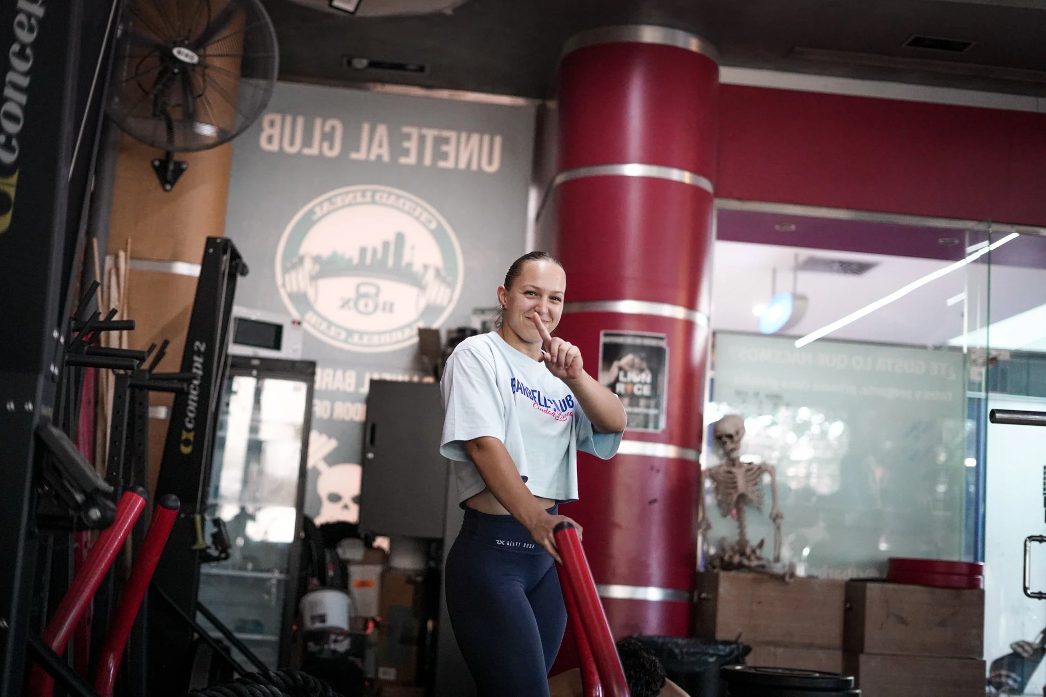 Una mujer en un gimnasio con un palo de entrenamiento, sonriendo y tocando su labio índice mientras mira hacia la cámara, con fondo de equipo de ejercicio, pared decorada y un esqueleto de gimnasio.