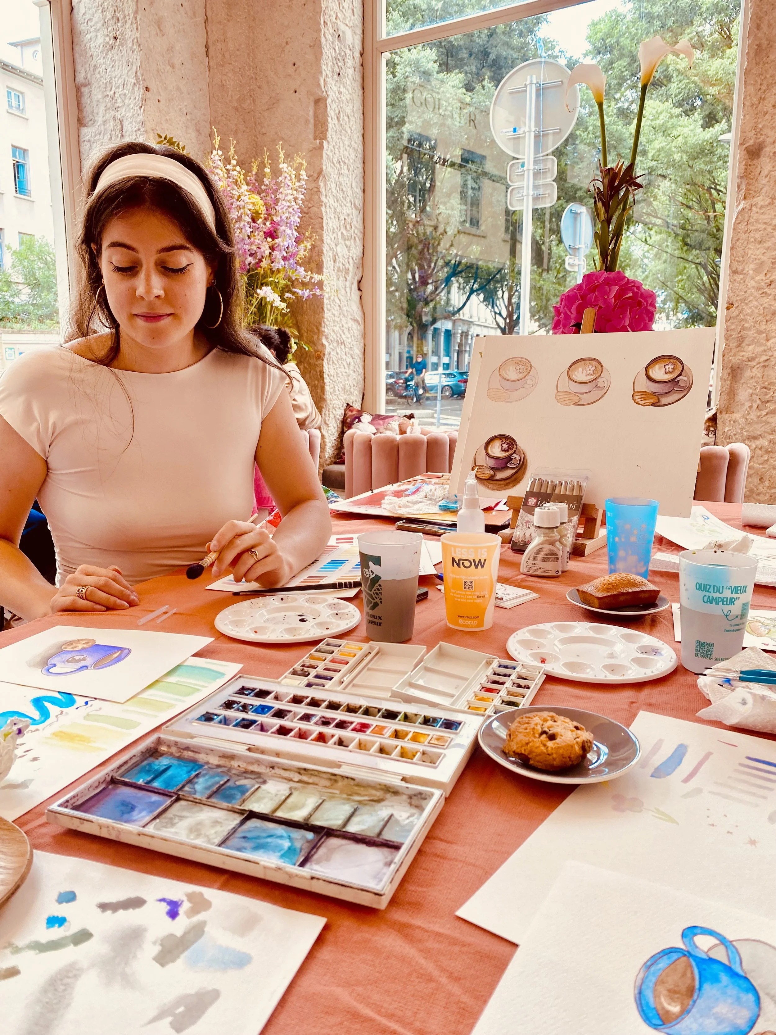 Une femme qui peint avec de l'aquarelle dans une ambiance lumineuse avec de la végétation visible à travers la fenêtre.