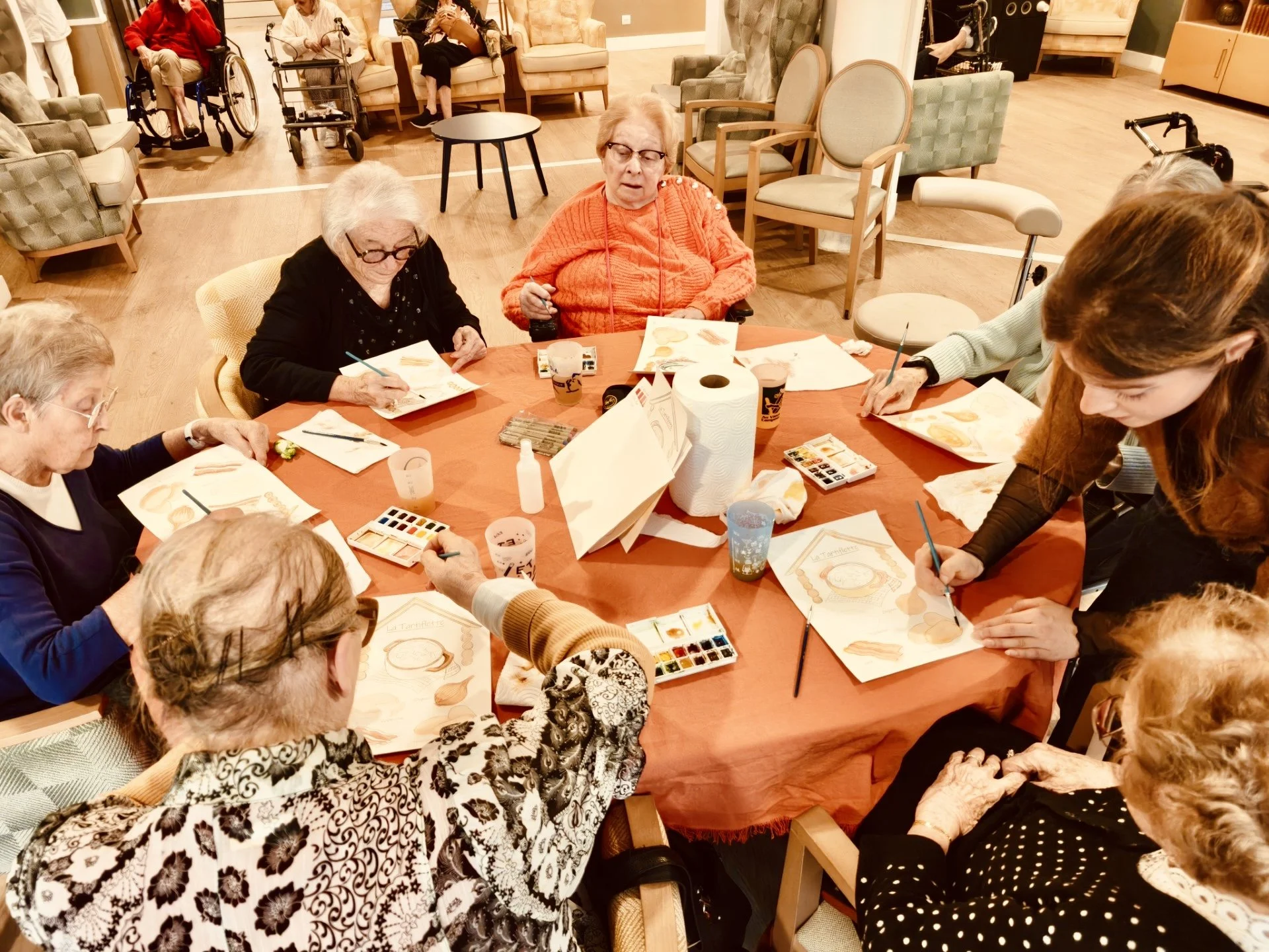 Groupe de personnes âgées assises autour d'une table, en train de peindre ou colorier. La scène se déroule dans une salle chaleureuse, probablement dans une maison de retraite ou un centre pour personnes âgées.