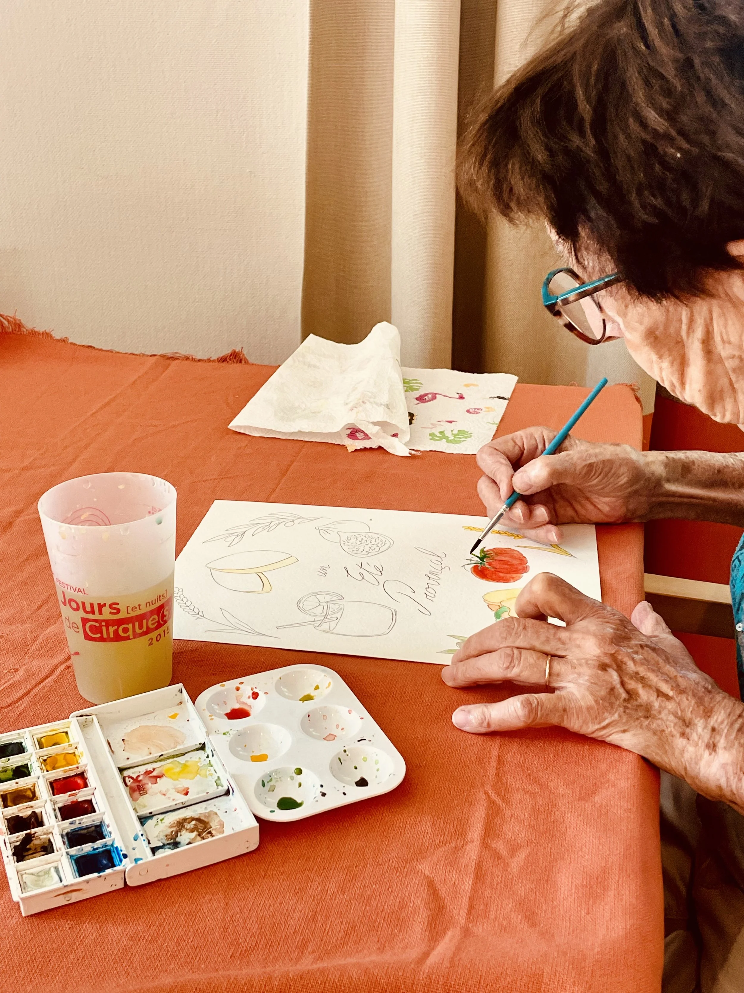 Une personne âgée qui peint une esquisse et ajoute de la couleur à un dessin avec de l'aquarelle sur une table recouverte d'une nappe. Il y a une palette de peinture, un verre et un papier avec des motifs de fleurs.
