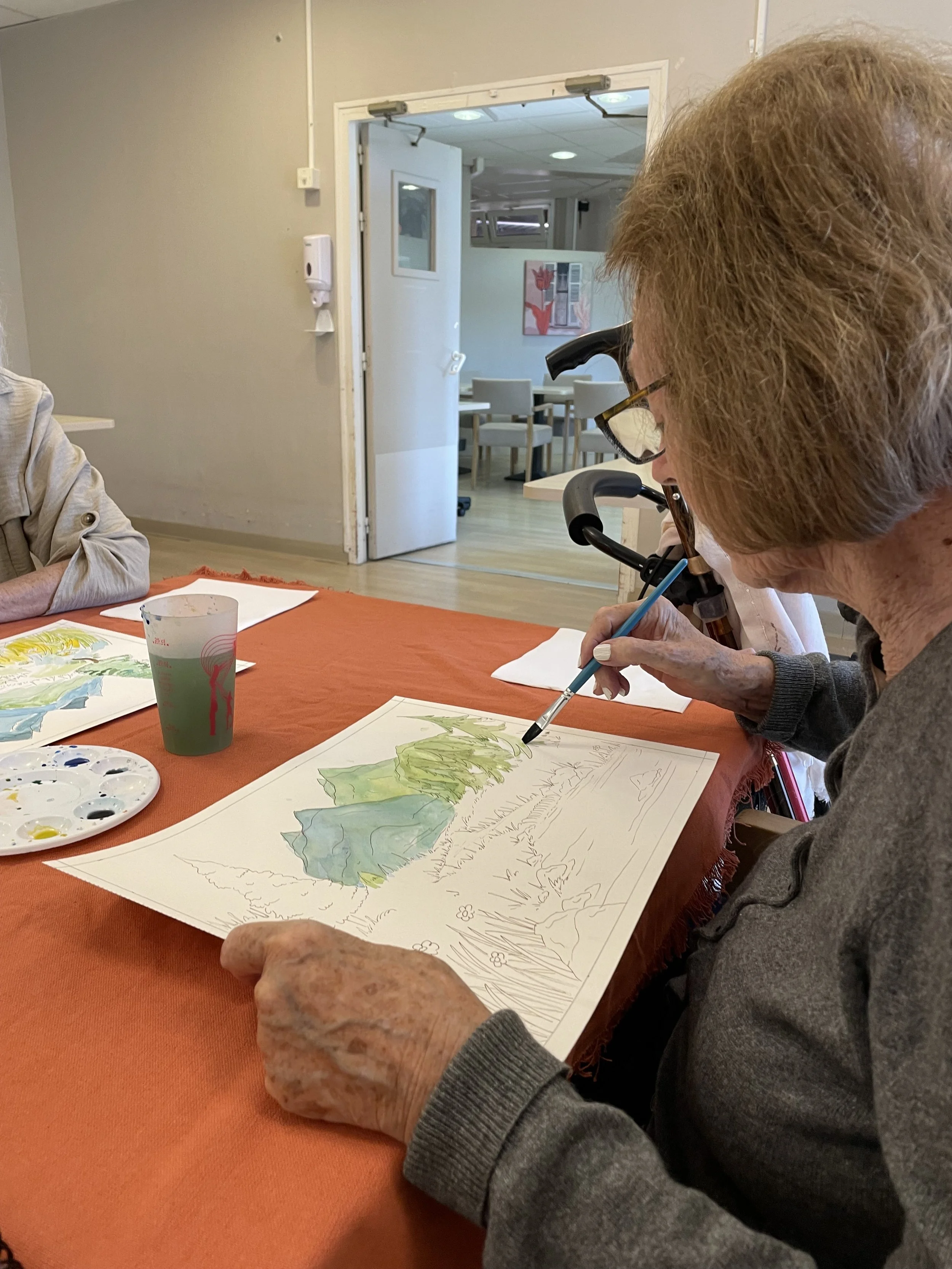 Une femme âgée en fauteuil roulant peint une toile à l'aquarelle représentant un paysage avec des montagnes vertes, en utilisant un pinceau, dans une salle avec une table orange, des papiers, un plateau de peinture et un verre à moitié rempli de liquide.