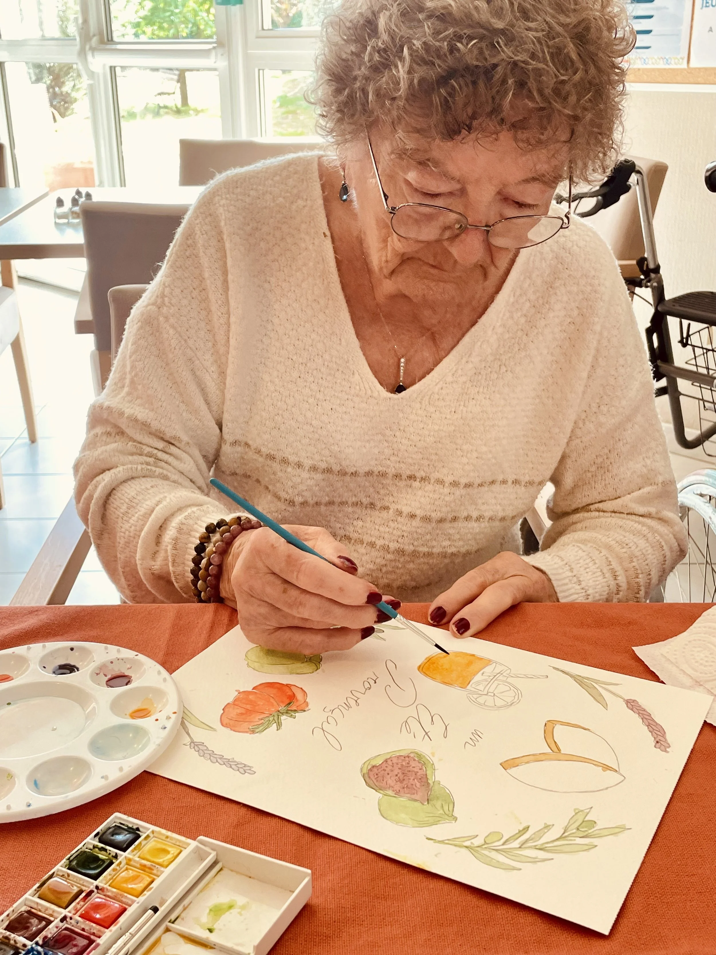 Une femme âgée fait de la peinture à l'aquarelle, représentant des légumes, dans une pièce lumineuse.