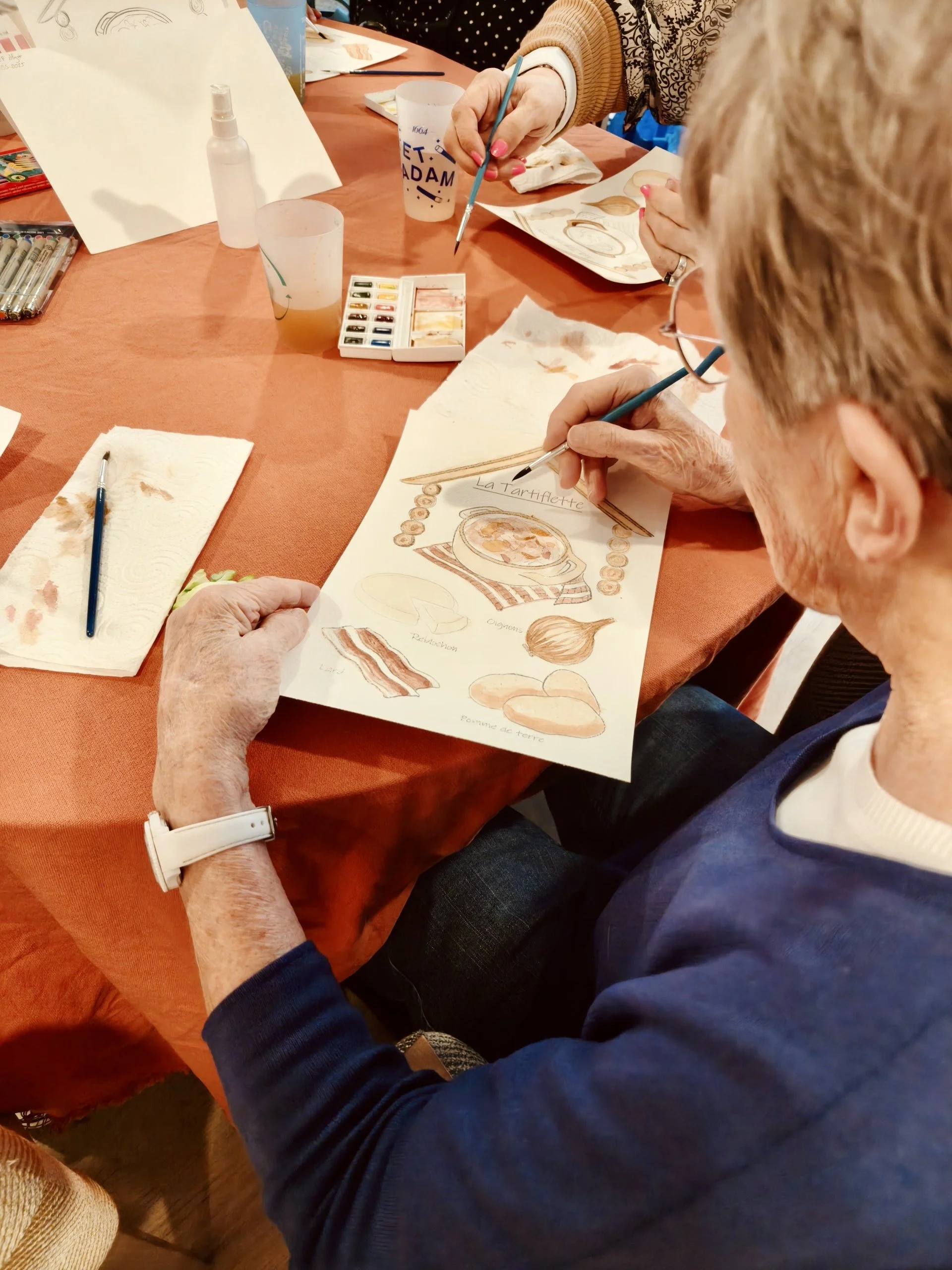 Une personne âgée peignant une illustration de la tarte Tatin avec des ingrédients comme la pâte, la pomme de terre, et l'oignon.