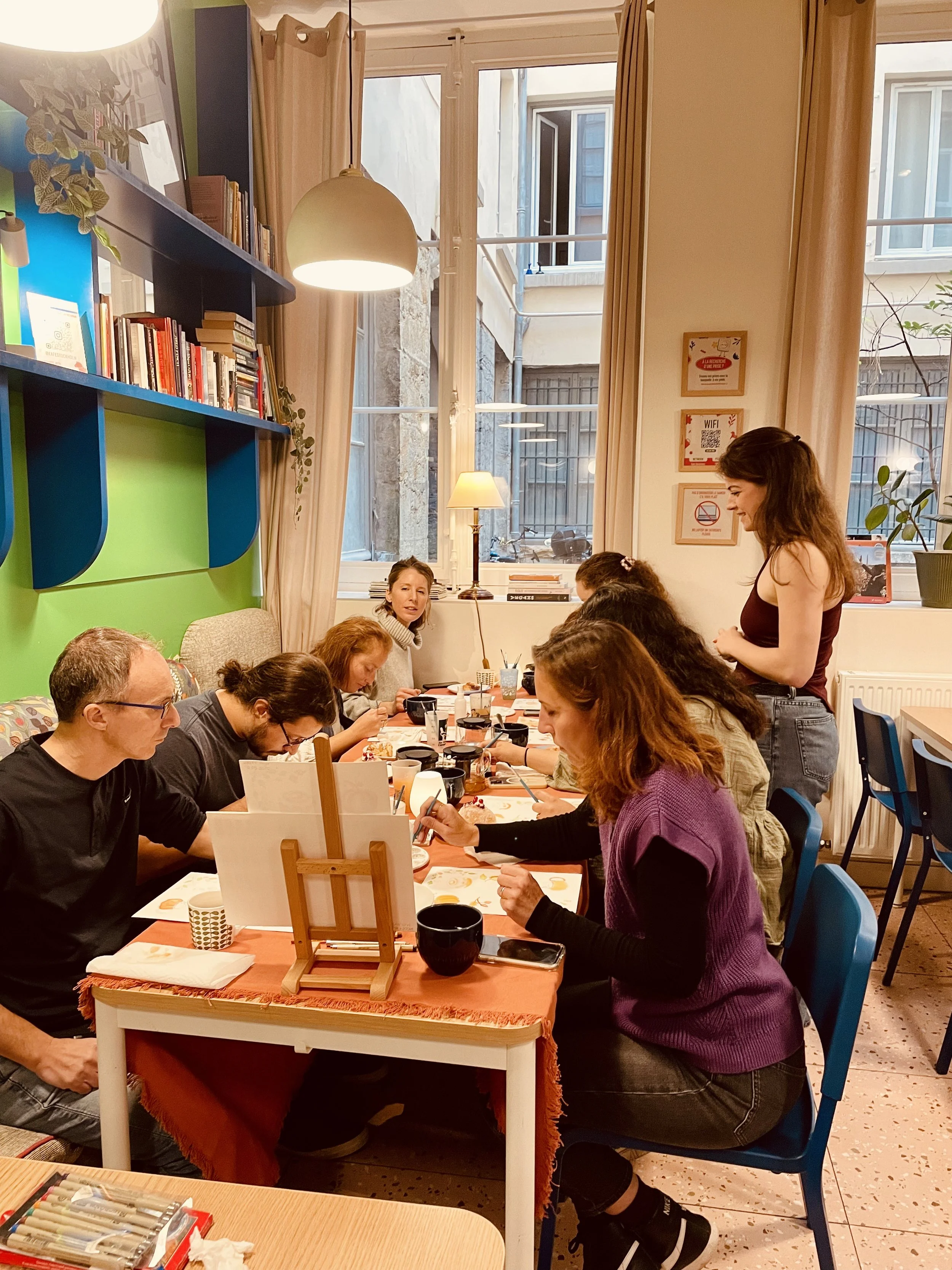 Groupe de personnes assises autour d'une table, en train de peindre dans une salle bien éclairée par une grande fenêtre, avec des livres sur une étagère et des décorations murales.