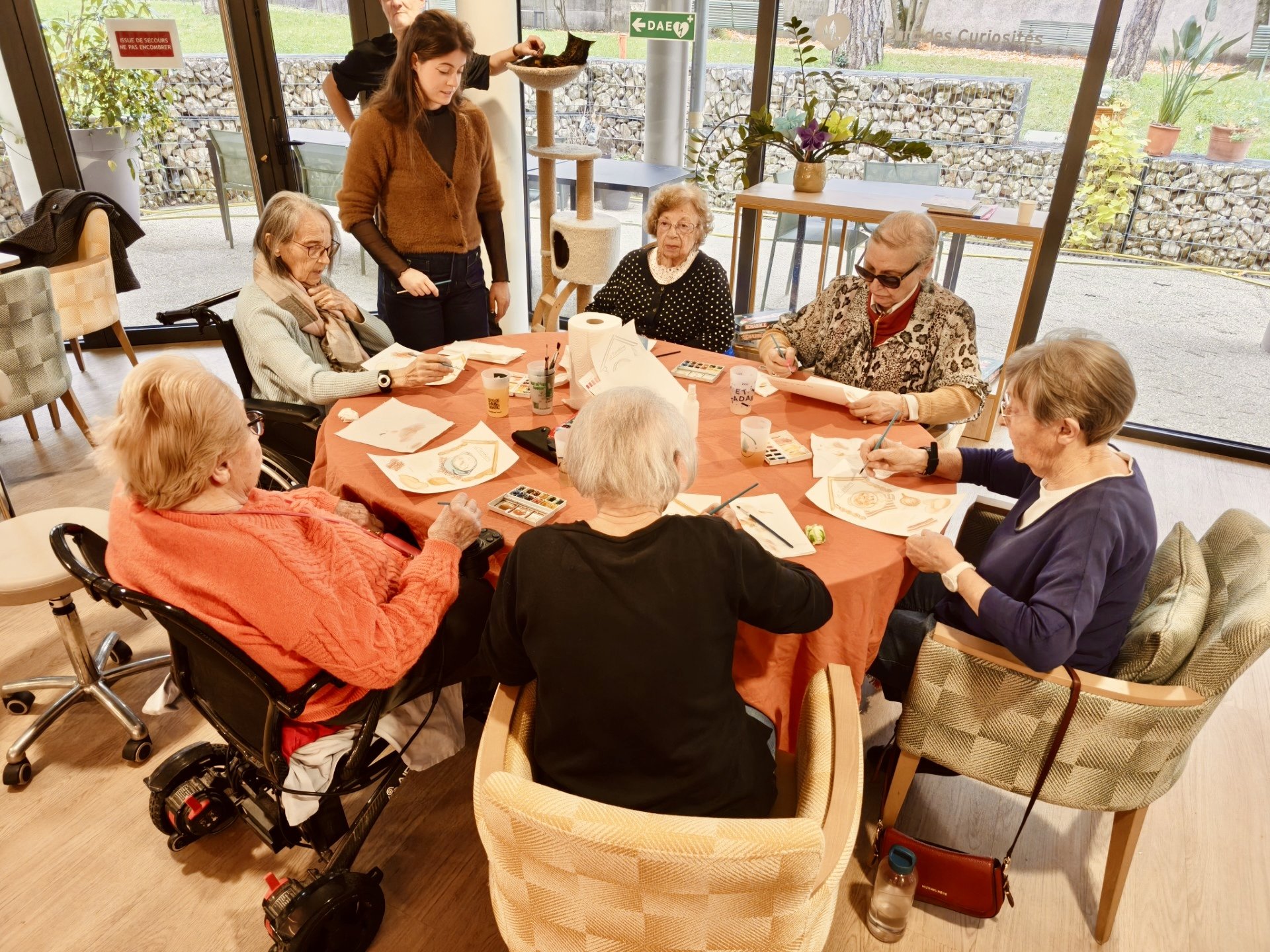 Groupe de personnes âgées assises autour d'une table, la plupart peignant avec des aquarelles, dans une pièce lumineuse avec de grandes fenêtres et un extérieur visible.