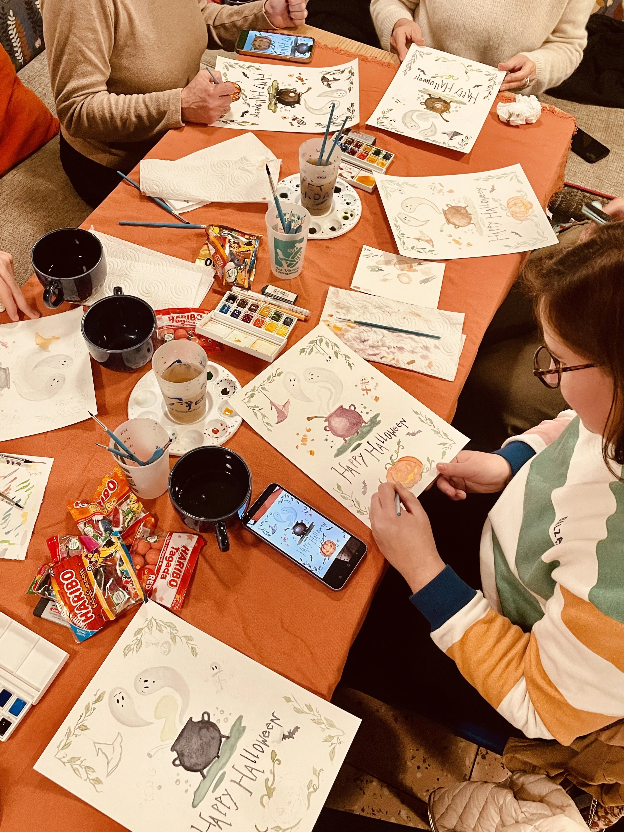 Groupe de personnes peignant des affiches de Halloween à une table, avec de la peinture, des pinceaux, des bonbons et des verres de boisson.