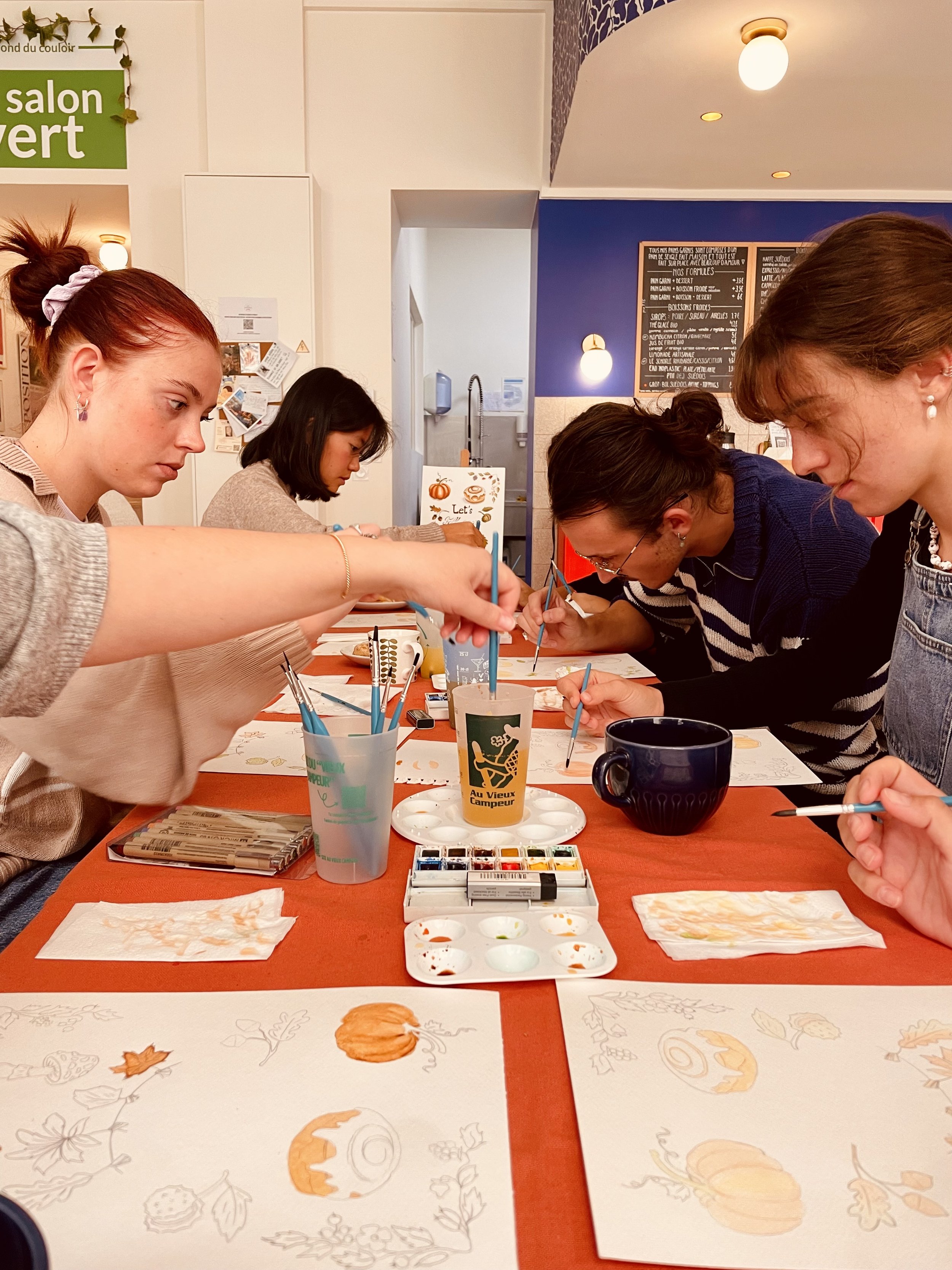 Quatre femmes participent à un atelier de peinture avec des motifs d'automne, tels que des citrouilles et des feuilles, sur la table. Elles utilisent des pinceaux et de la peinture pour décorer des feuilles de papier ou des cartes.