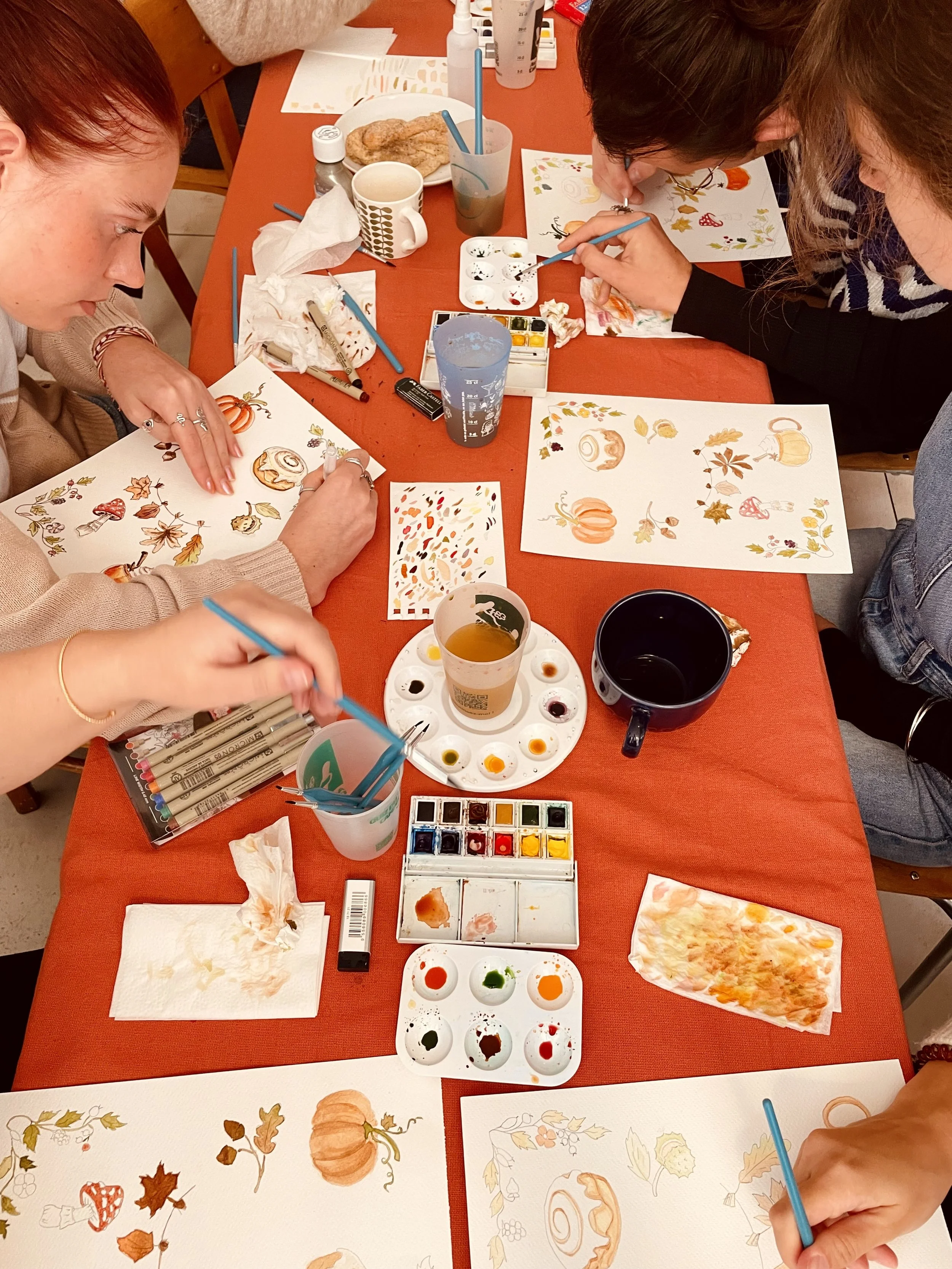 Quatre personnes font de la peinture sur des feuilles blanches avec des motifs d'automne, tels que des citrouilles, des feuilles et des champignons. La table est équipée de petits pots de peinture, de pinceaux, d'une palette, de boissons et de papiers absorbants.