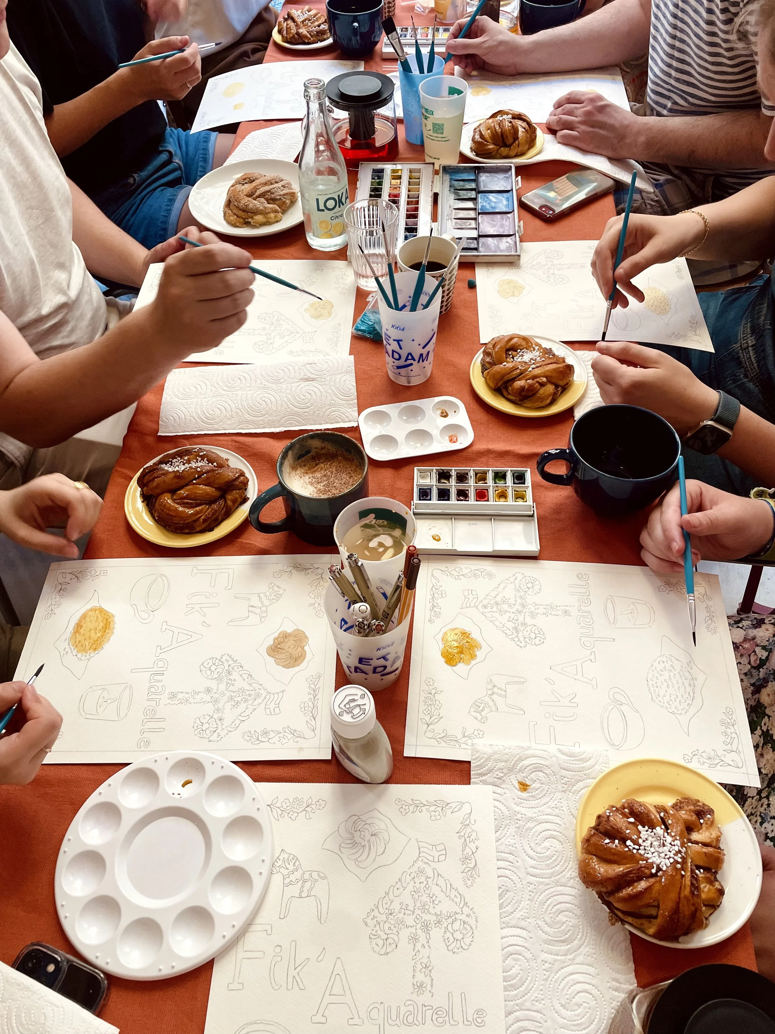 Groupe de personnes en train de peindre des dessins sur des feuilles blanches avec des pinceaux, entouré de pâtisseries, de tasses, d'une palette de peinture et d'eau, sur une table avec un napperon orange.