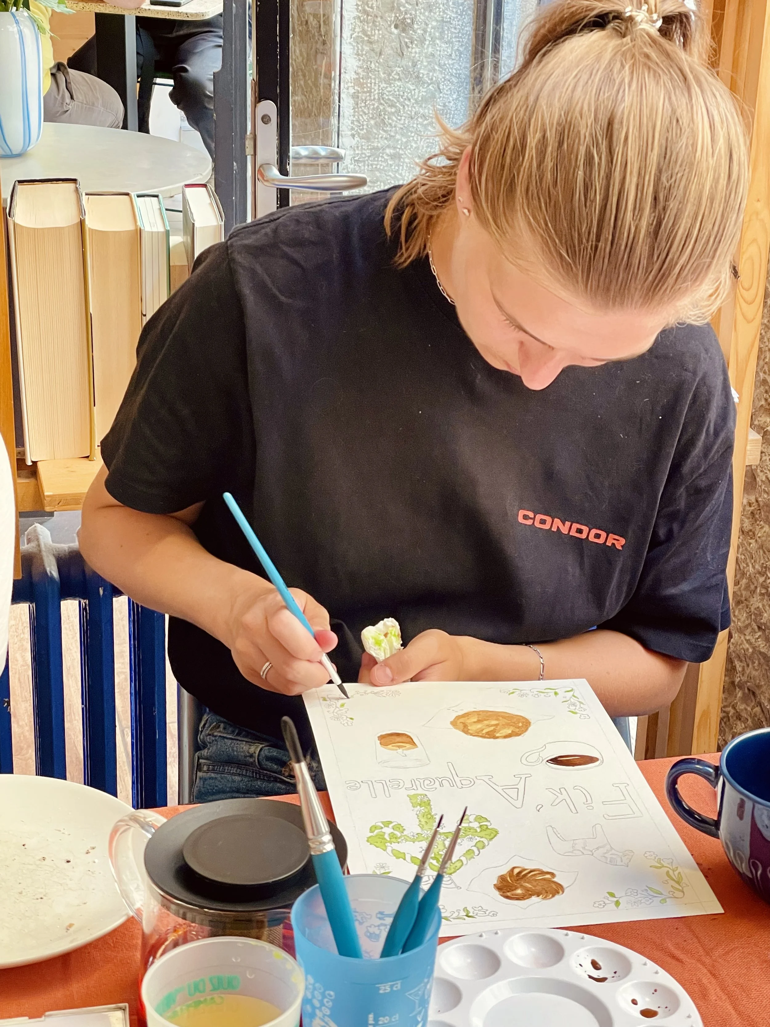 Une femme peint un poster avec diverses illustrations et textes à l'intérieur d'une pièce lumineuse, assise à une table avec des fournitures d'art et un mug.