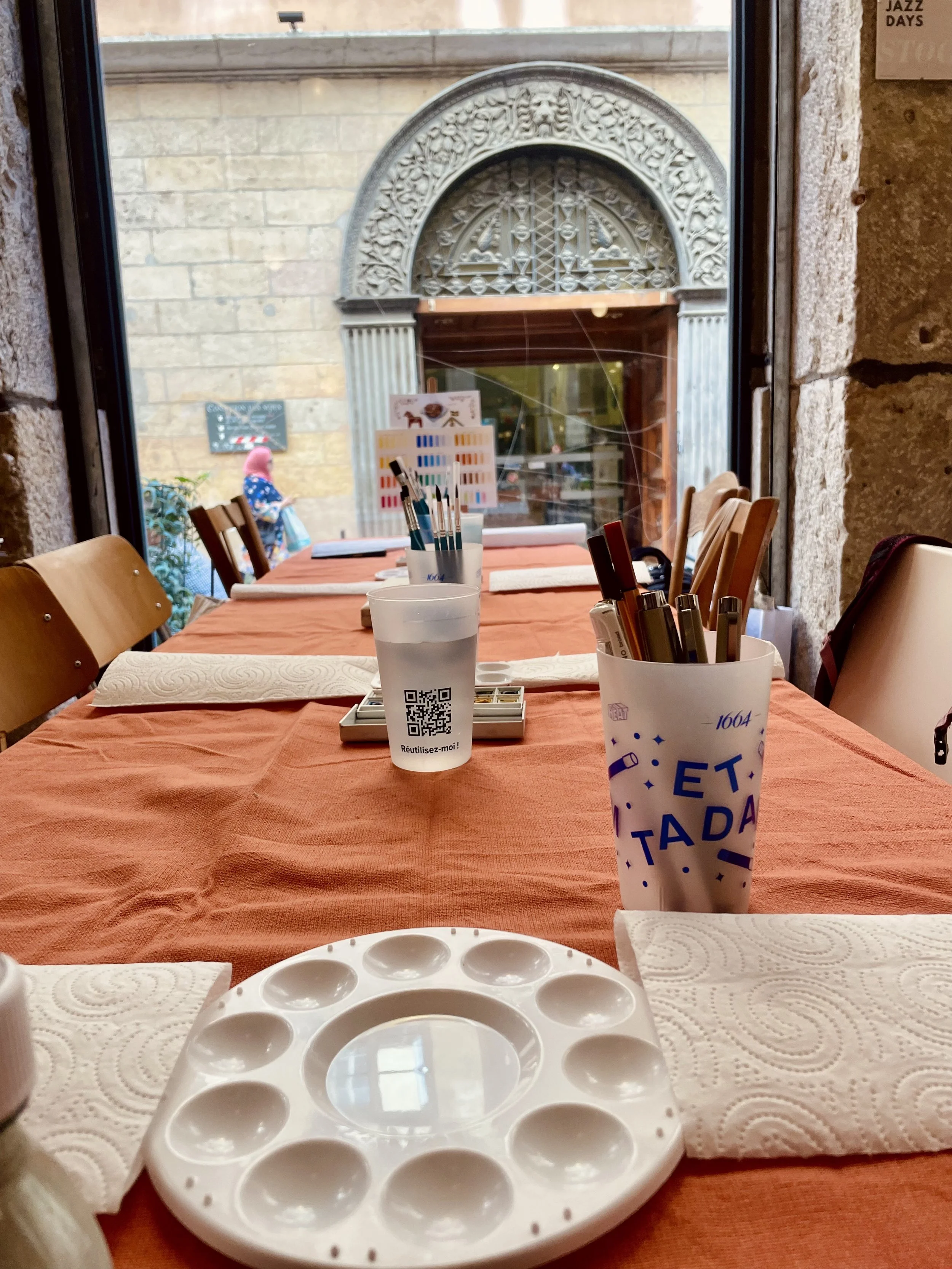 Une table de peinture avec un plateau, des pinceaux, un verre avec une QR code, un porte-stylo avec des marqueurs, une serviette en papier, et une nappe orange, face à une grande fenêtre avec vue sur la rue et un bâtiment en pierre avec une arche en pierre ornée des détails sculptés.