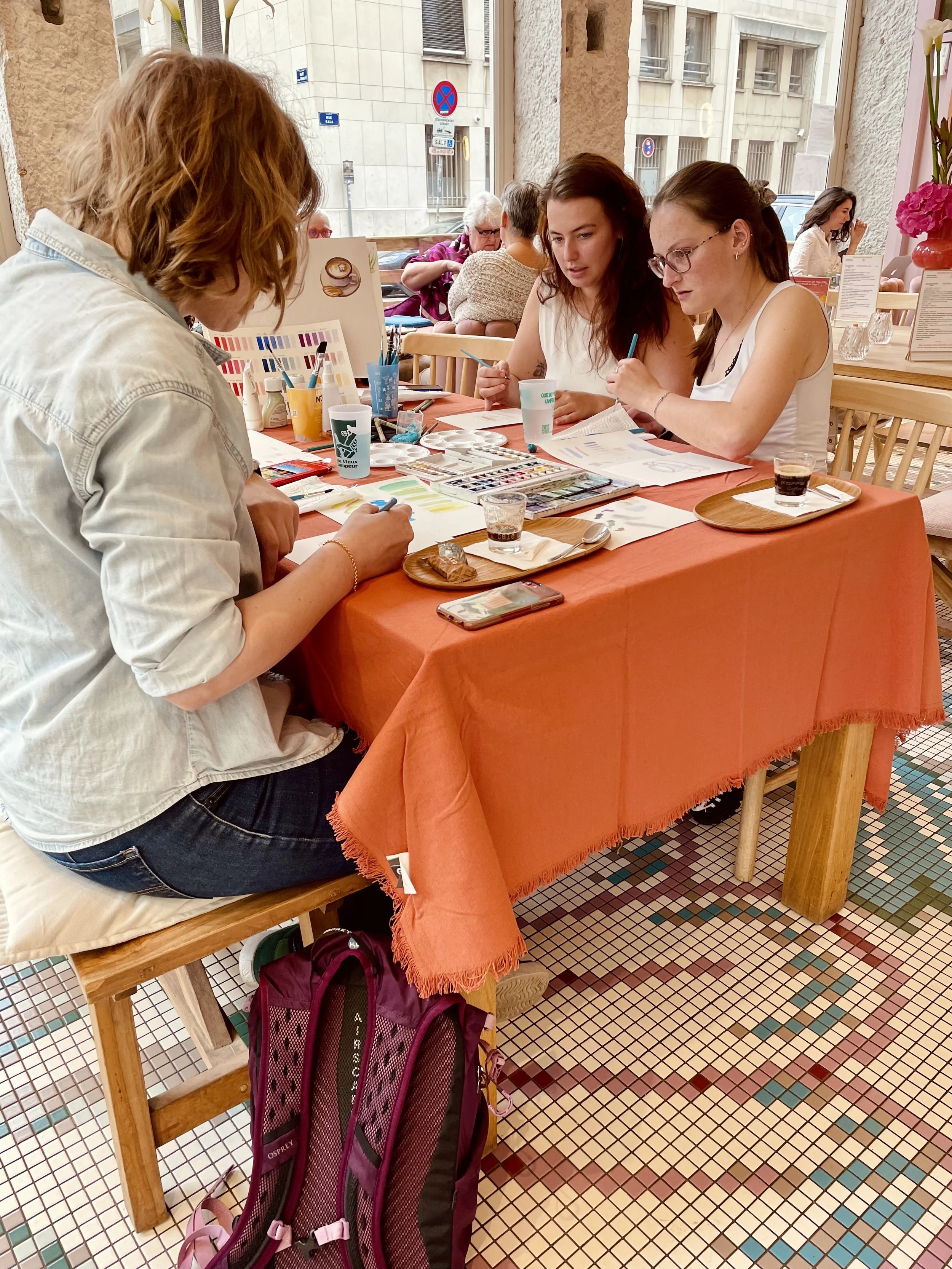 Trois femmes assises autour d'une table orange en train de peindre ou dessiner, dans un café ou restaurant. La table est couverte de matériel artistique, contenant des pinceaux, palette de couleurs, et autres outils. Il y a des verres de boisson sur la table.