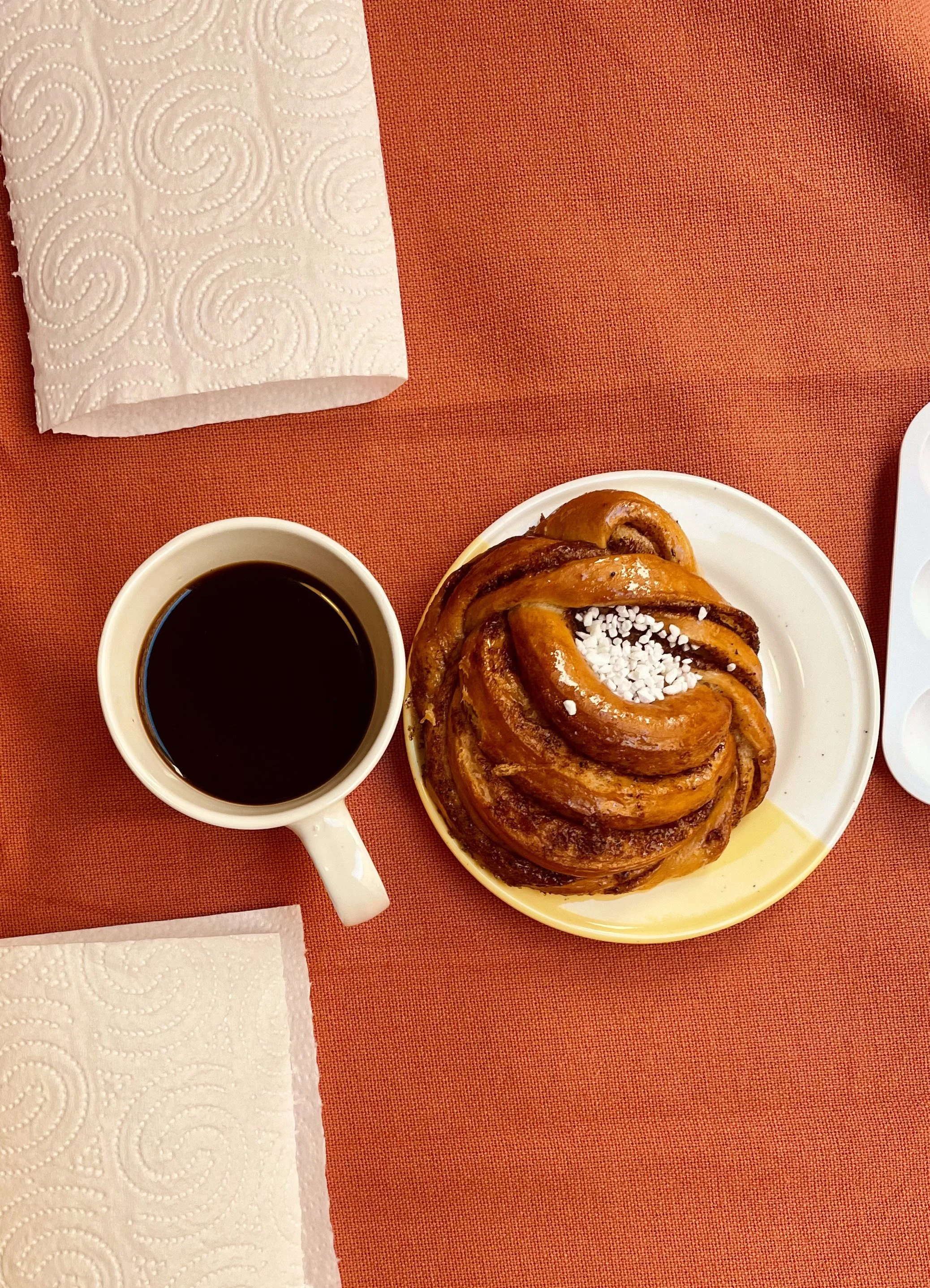 Un café noir dans une tasse blanche, une brioche tressée avec du sucre en grains sur une assiette blanche, posés sur une nappe rouge.