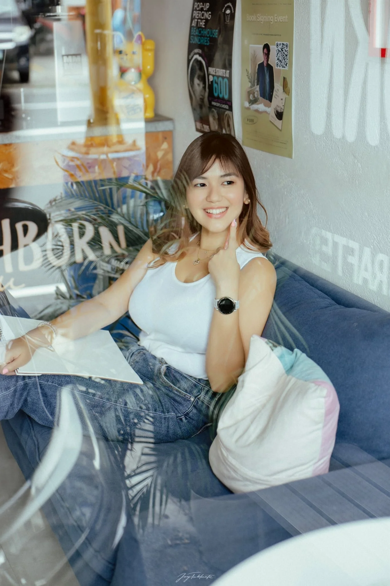 A smiling woman with brown hair sits on a blue couch inside a cafe, looking through a window at street scene. She wears a white tank top, jeans, a black watch, and a heart-shaped necklace. There are posters and a plant visible on the wall behind her.