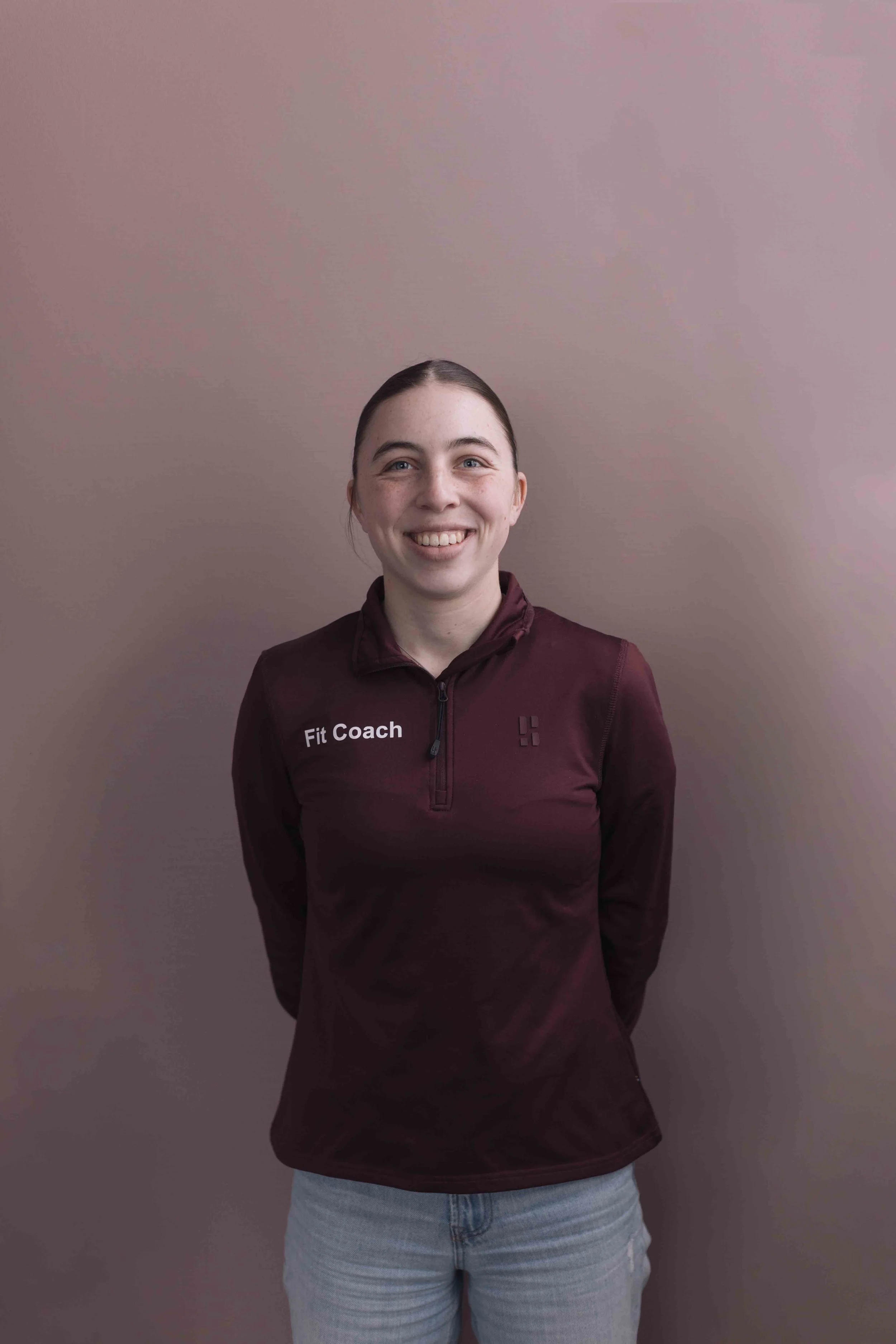 Britt Epping Goed Fysio Een jonge vrouw met bruin haar en een brede glimlach, gekleed in een donkermaroon vest met 'Fit Coach' tekst, staat voor een effen, lichtbruine muur.