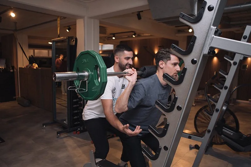 Twee mannen oefenen samen met gewichtheffen in een sportschool. Eén man helpt de ander die een barbell met gewichten afwisselend op en neer tilt.