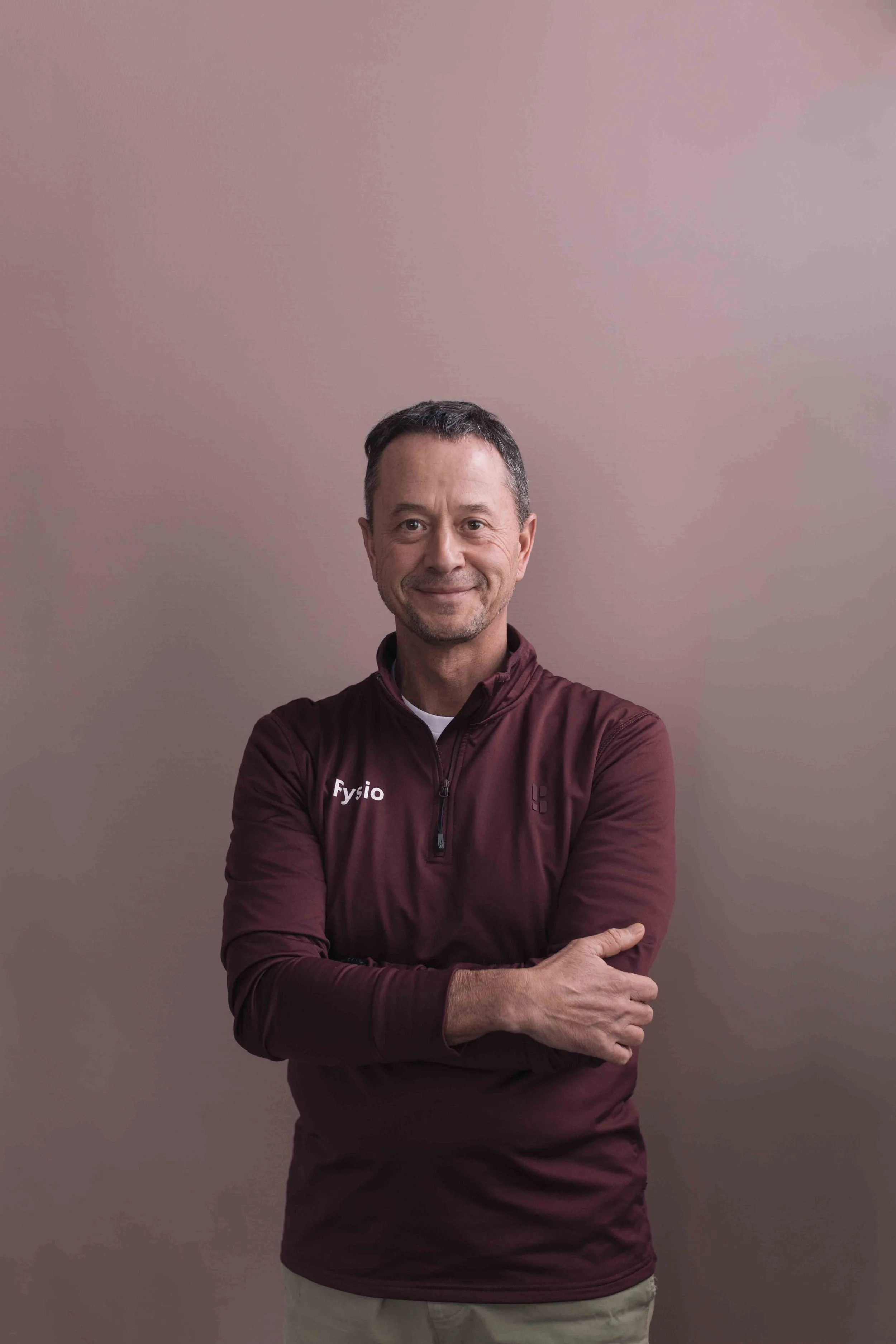 Man in burgundy sportsjack met 'Fysio' op de linkerborst staand met gekruiste armen voor een beige muur.