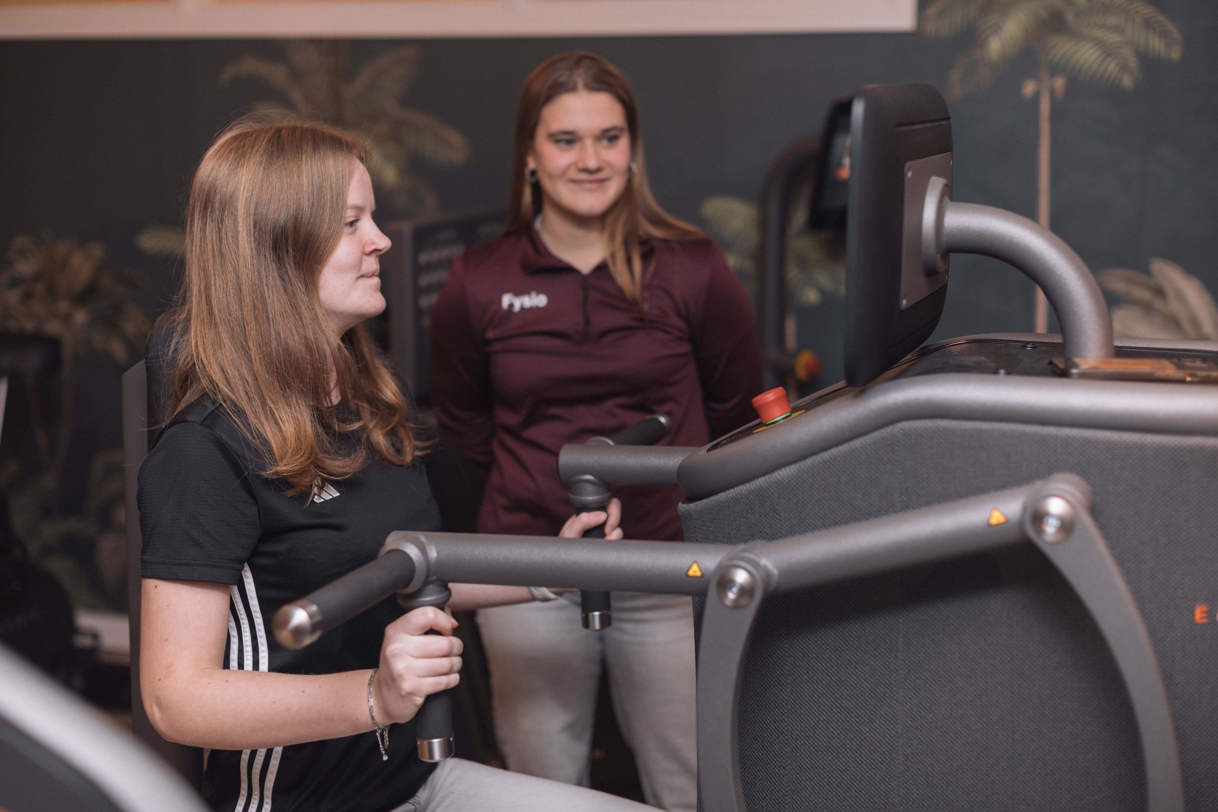 Goed Fysio Twee vrouwen in sportschool, een aan het joggen op loopband en de ander kijkt toe.