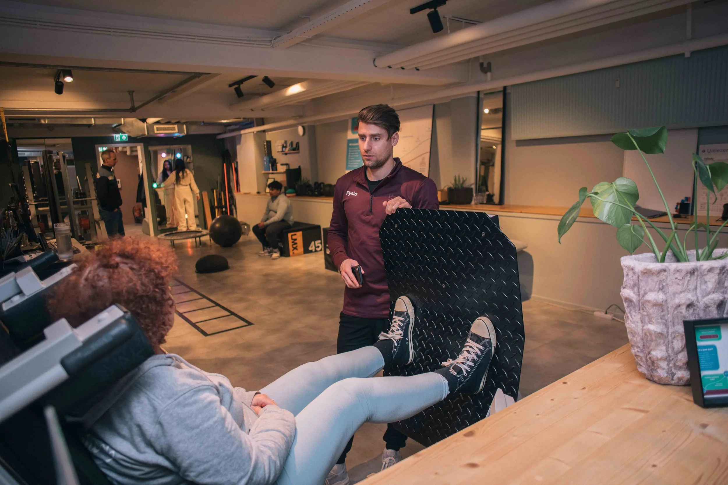 Een fitnessruimte met verschillende mensen die fitnessactiviteiten doen. Een man in sportkleding staat met een voet op een bankje, terwijl een persoon met witte sportkleding met benen omhoog ligt op een andere bank. Anderen bewegen in de achtergrond.