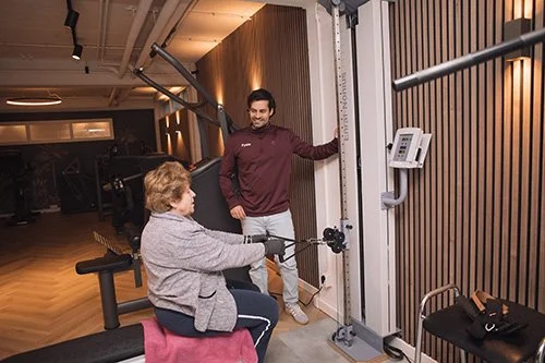 Een man begeleidt een vrouw die een lichte beperking heeft bij het gebruik van een fitnessapparaat in een sportschool.