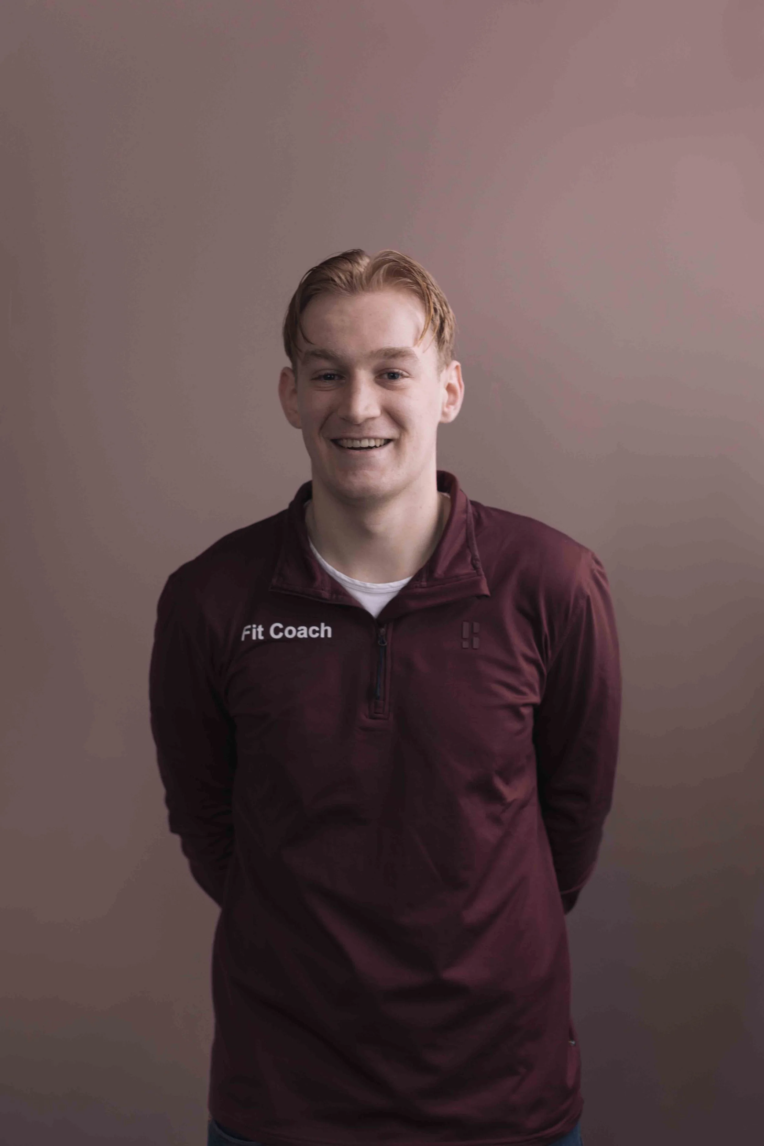 Joris Zandbergen Goed Fysio een jonge man met rood haar, lachend, draagt een burgundy trainingsjack met de tekst 'Fit Coach' op de borst, staat voor een effen grijze achtergrond.