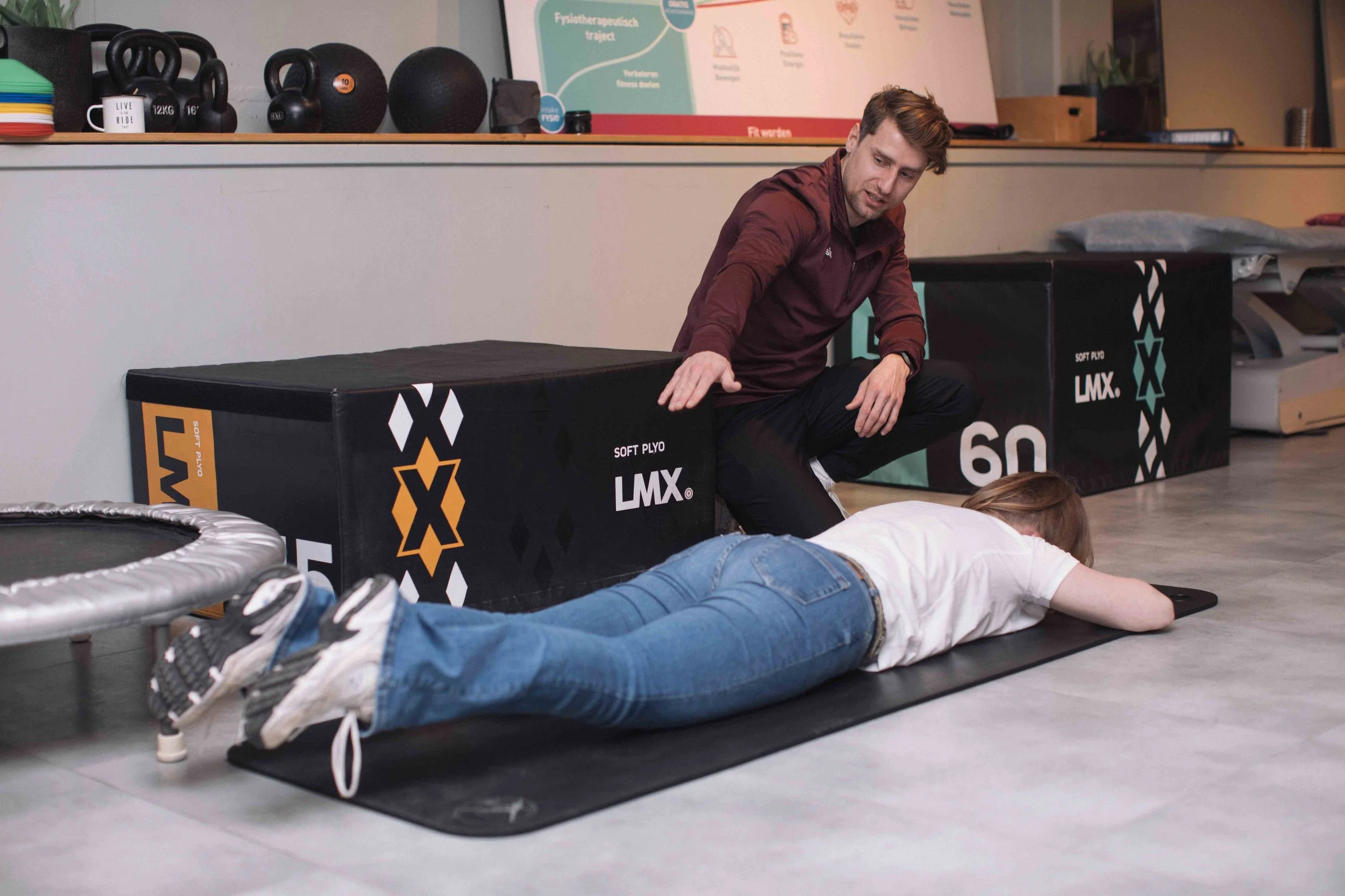 Goed Fysio Een man in een sportzaal geeft een eerste hulp cursusbijeenkomst en helpt bij het reanimeren van een vrouw die op haar buik op de grond ligt. Op de achtergrond bevinden zich fitnessapparatuur en een bord met informatie.