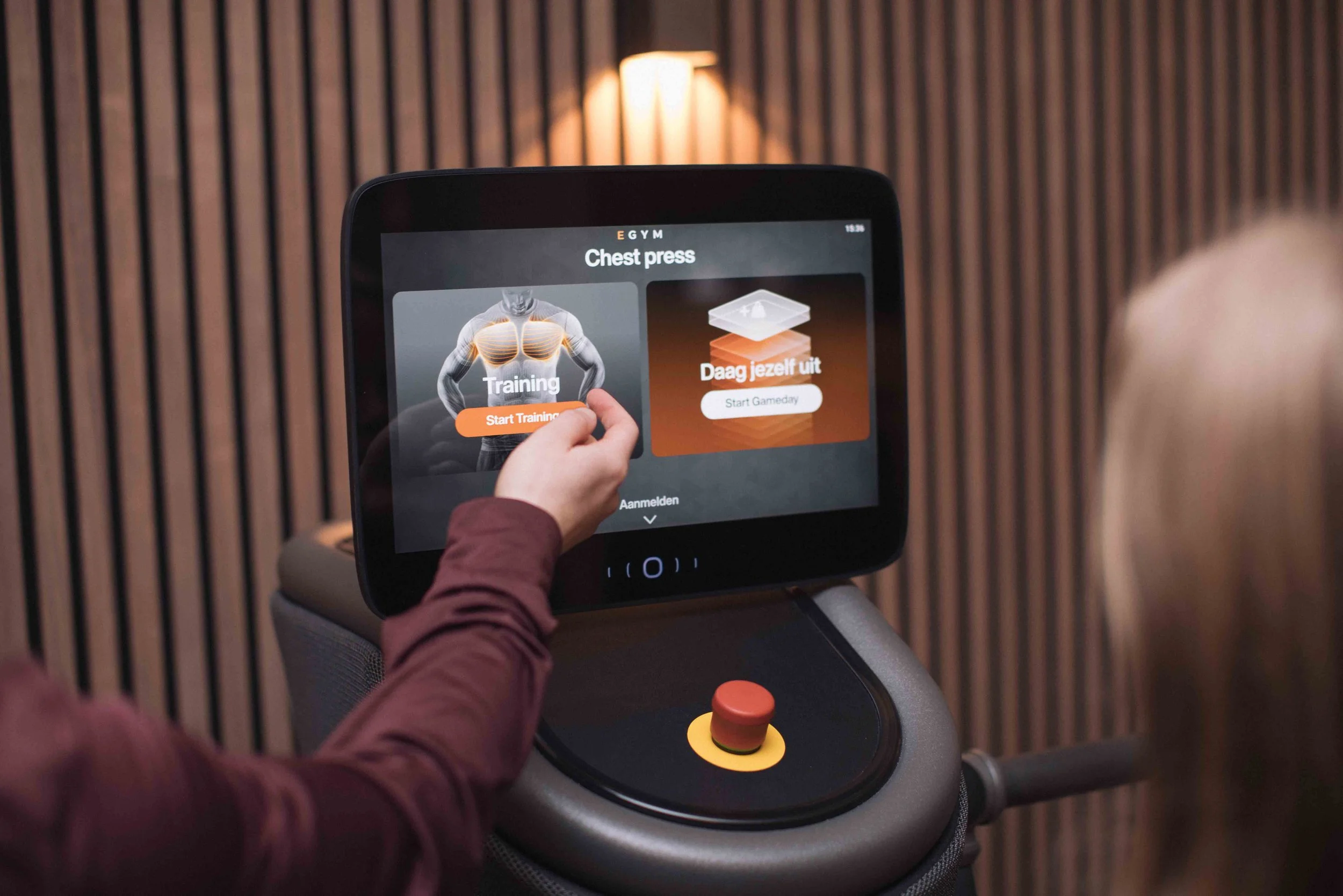 Persoon bedient een fitnessapparaat met een scherm waarop oefeningen worden weergegeven, in een ruimte met houten wand.