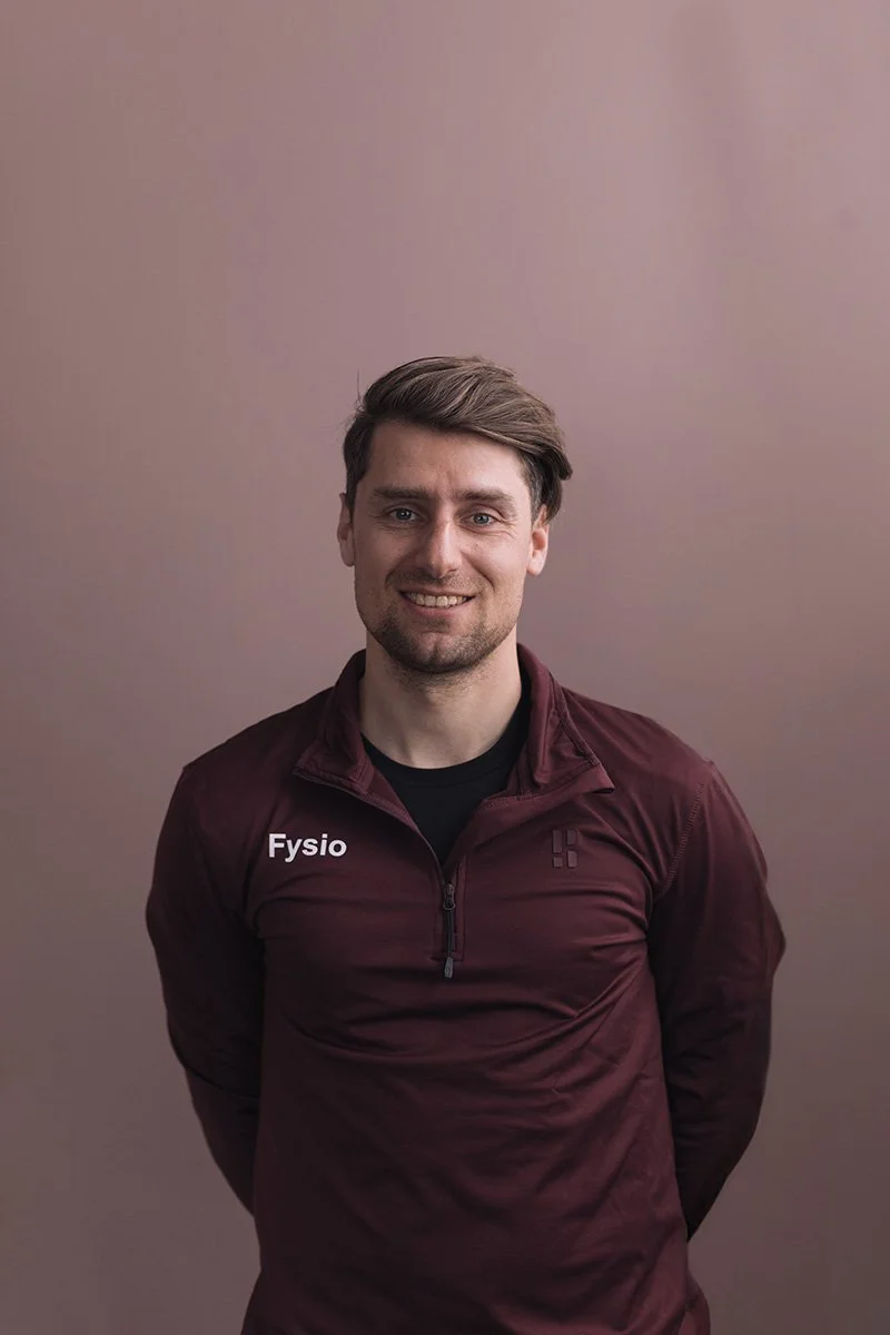 Mark Bukman Goed Fysio Man in een bruin trainingsjack met het woord 'Fysio' op de linkerborst, lachend, in voorgrond, met een effen beige muur op de achtergrond.