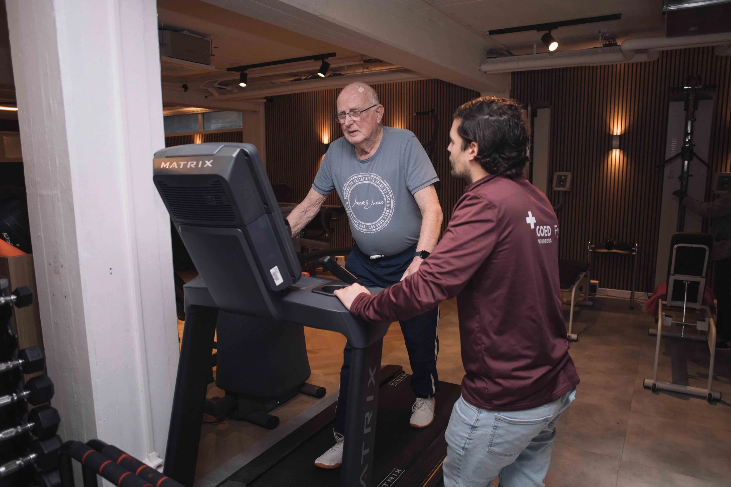 Goed Fysio Een oude man en een jonge man trainen op loopbanden in een sportschool, met houten wandpanelen en fitnessapparatuur op de achtergrond.