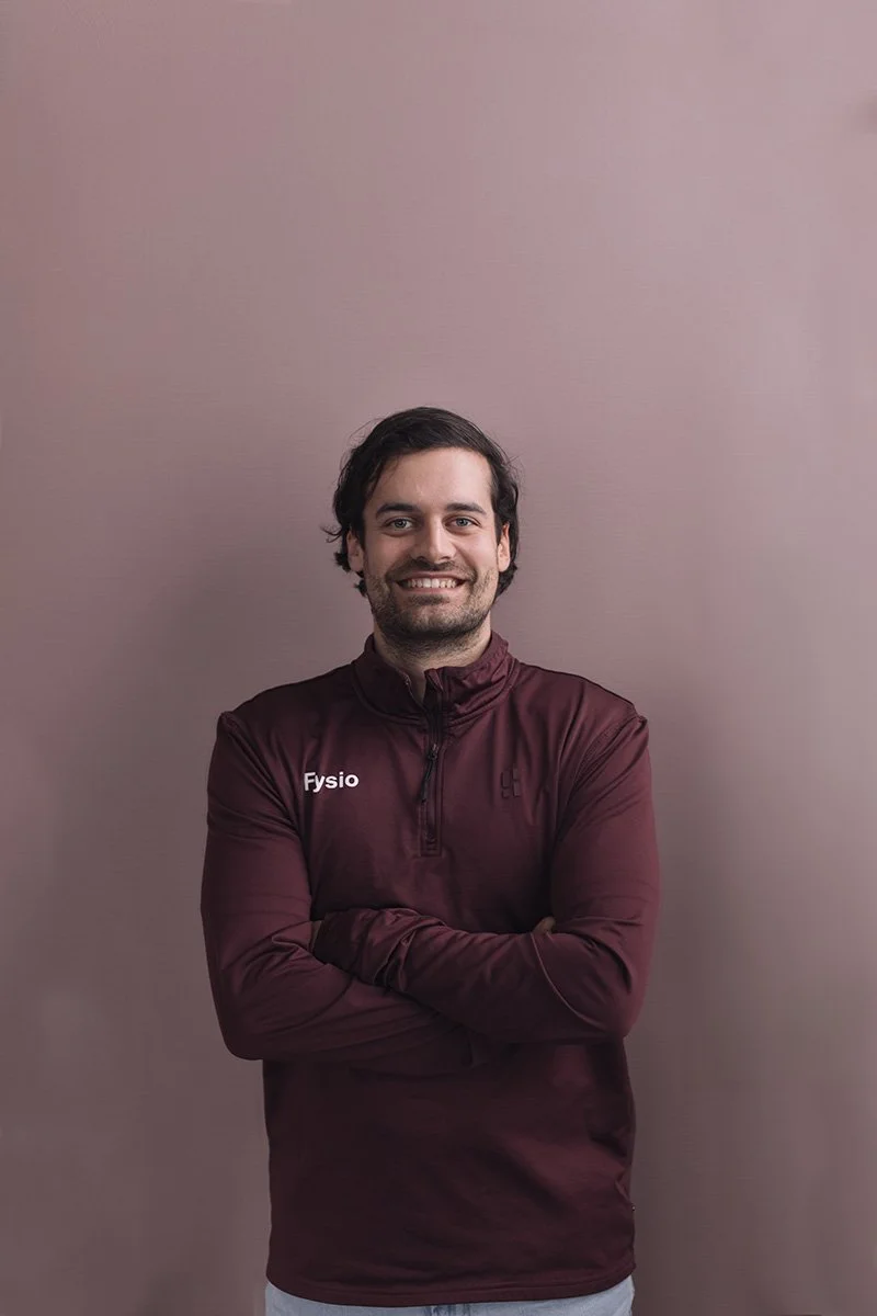 Bas van de Velde Goed Fysio Een man met donker haar en een gezichtsbeharing, lachend, gekleed in een donkerrode sportjas, staand tegen een effen lichtpaarse achtergrond.