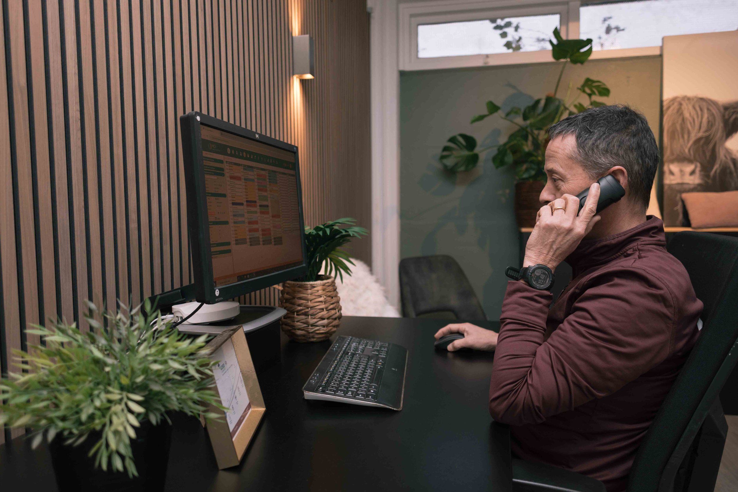Goed Fysio Man in kantoor met computer en telefoon, zittend aan bureau met planten en houten wand.