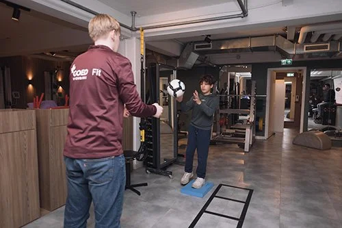 Een persoon traint met een fitnesscoach in een sportschool, oefent met een medicijnbal, terwijl een ander kind op een balanceermat staat.