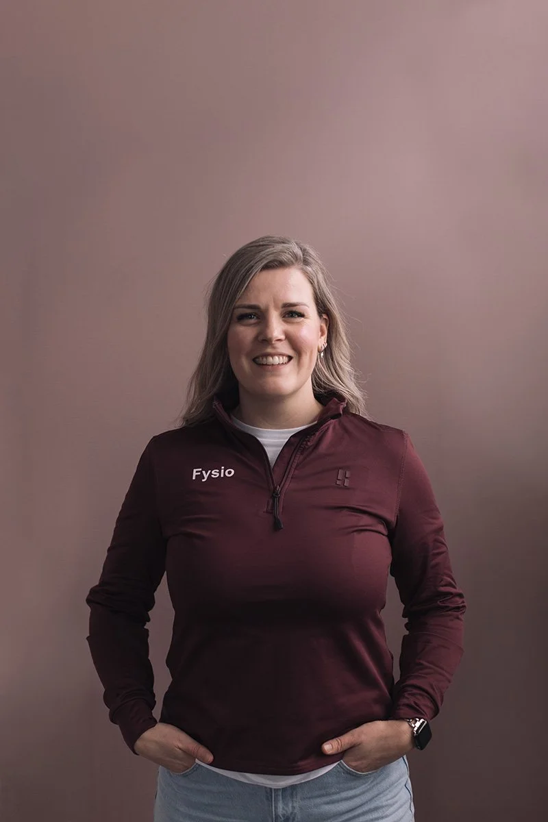 Suzanne de Jong Goed Fysio Vrouw staat rechtop tegen een effen bruinachtige achtergrond, draagt een donkerrode sportjas met 'Fysio' op de borst, lachend met haar handen in haar zakken.