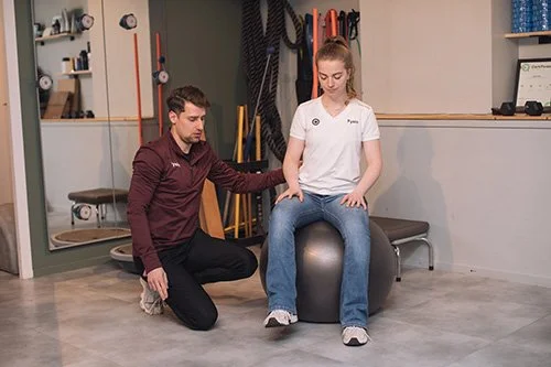 Behandelaar die een vrouw helpt met oefeningen op een fitnessbal in een fysiotherapiepraktijk.
