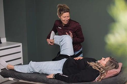 Een fysiotherapeut helpt een patiënt met een oefening, liggend op een behandelbank in een kliniek of revalidatiecentrum.
