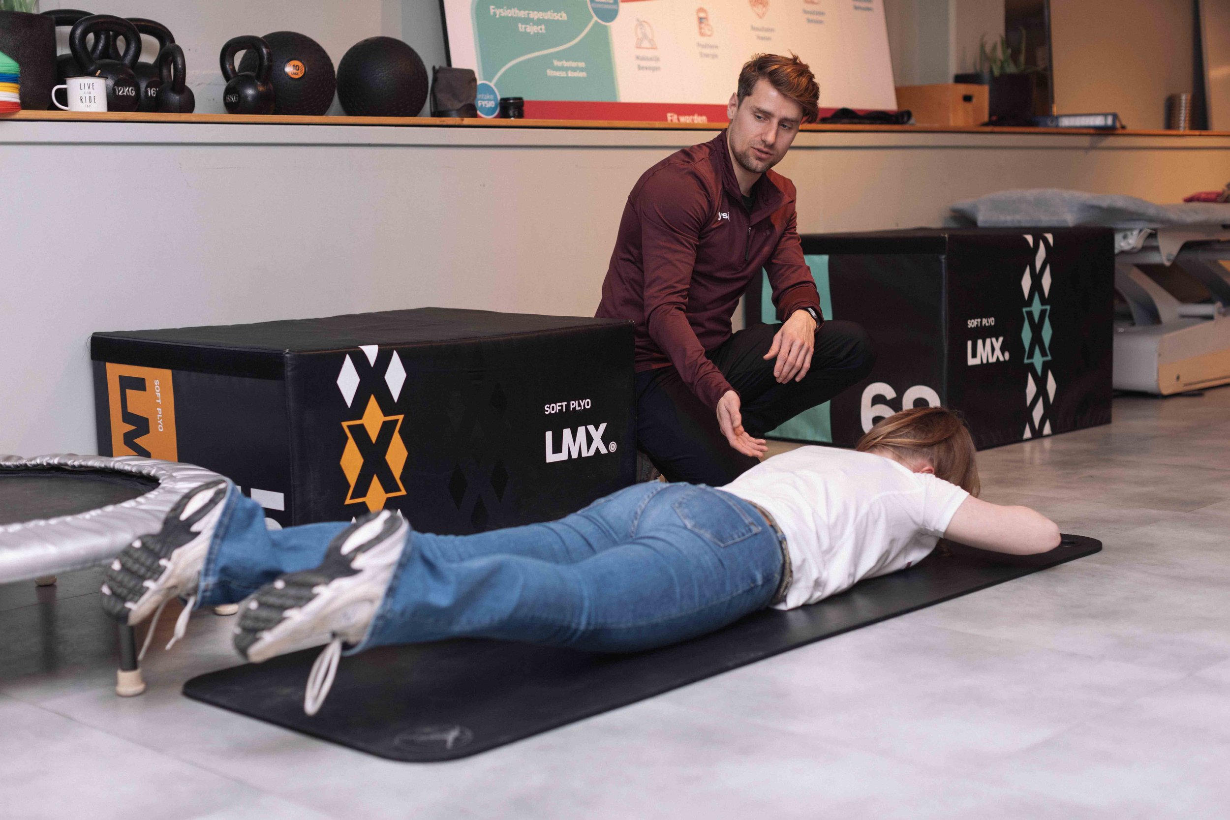 Goed FysioEen man die zich in een sportschool voorbereidt op een oefening terwijl een vrouw op de grond ligt, beiden op sportmatten. Op de achtergrond zijn gewichtskettingen, zwart speelgoed en een diagram te zien, wat wijst op een trainingsomgeving.