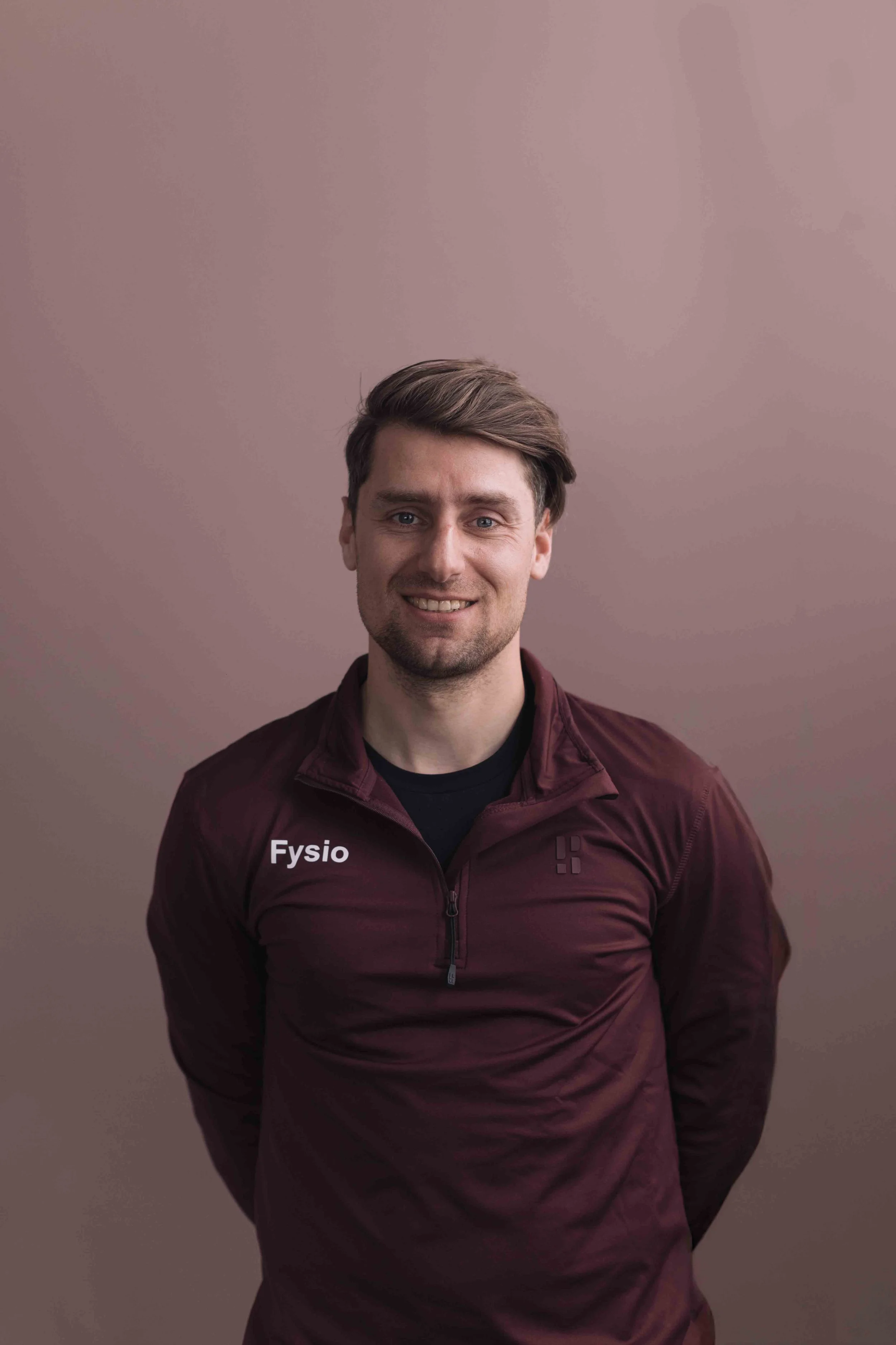 Een jonge man met lichtbruin haar en een glimlach, gekleed in een donkerrode sportjas met 'Fysio' erop, staat voor een effen, licht roze muur.